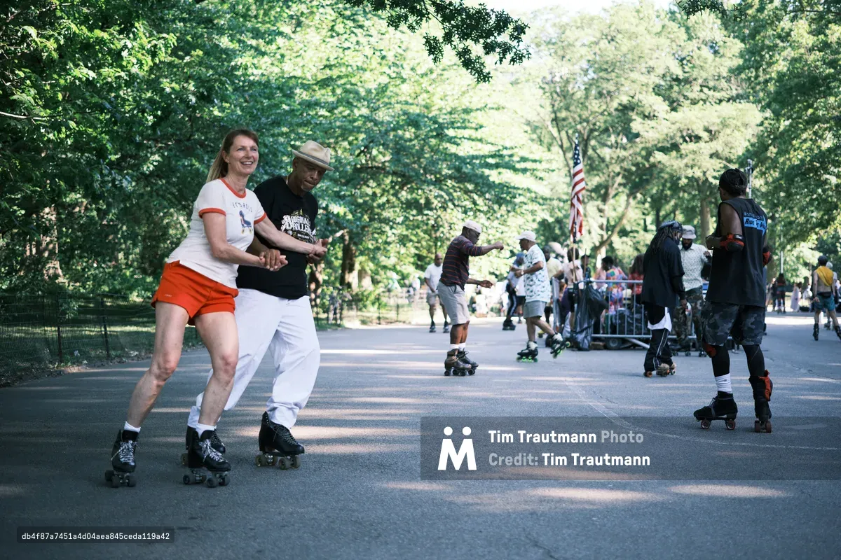 Two friends share a moment of pure delight as they roller skate together down a tree-lined pathway in Central Park. The woman in orange shorts and white shirt beams with laughter while holding hands with her companion in a beige hat and dark shirt, their synchronized movement captured against the dappled sunlight filtering through the lush summer canopy. Behind them, other skaters glide along the same path, creating a scene of community recreation beneath the park's towering trees. The warm, golden light and relaxed atmosphere perfectly capture the timeless appeal of roller skating culture in Manhattan's green heart.