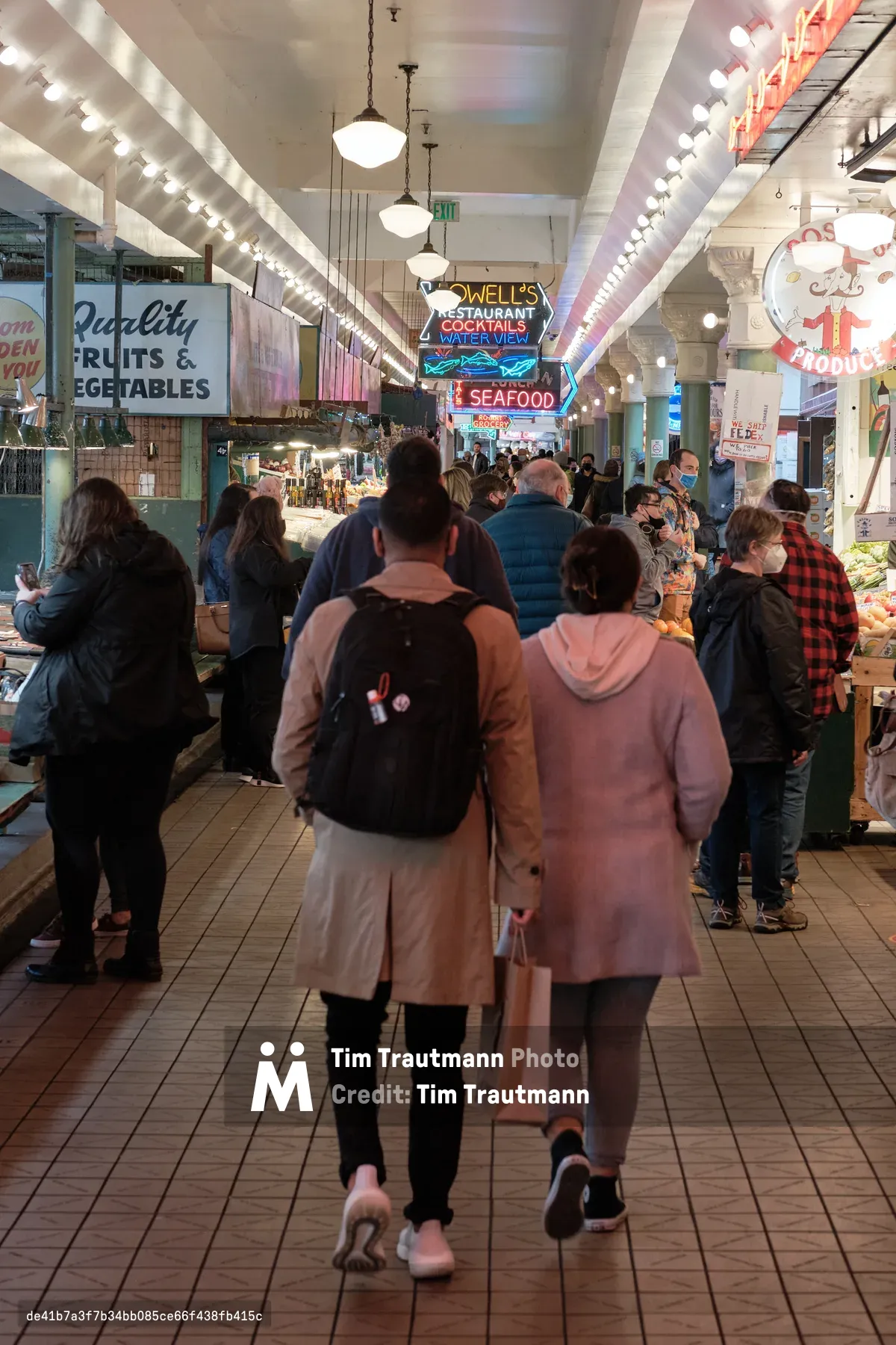 Crowds of visitors navigate the iconic main corridor of Pike Place Market, Seattle's historic public market. The scene captures the market's characteristic energy as shoppers meander past vintage neon signs, including Lowell's Restaurant and quality produce stalls. Overhead, strings of bright bulbs illuminate the white-painted steel beams and brick facades, creating the warm, inviting atmosphere that has drawn locals and tourists for over a century.