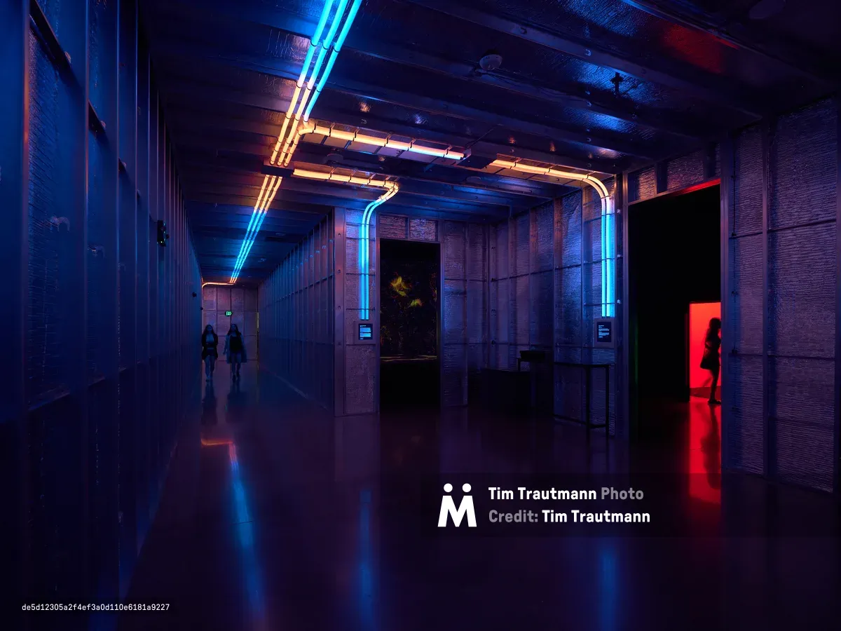 A dramatic interior corridor featuring striking blue and orange neon lighting strips along the ceiling and walls, creating a cyberpunk atmosphere with silhouetted figures walking through the illuminated space. The modern architectural design includes exposed ceiling elements and reflective flooring that amplifies the colorful LED lighting effects.