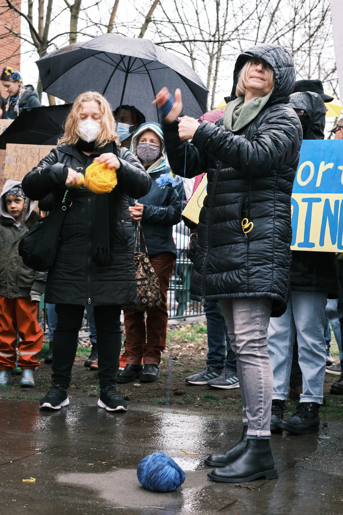 Bundled against Portland's winter chill, protesters gather outside Revolution Hall in the Central Eastside, their breath visible in the cold air as they demonstrate against the looming threat of war in Ukraine. Rain-slicked pavement reflects the gray afternoon light while masked demonstrators clutch signs and umbrellas, embodying both vulnerability and determination. The scene captures the prescient urgency of citizens mobilizing days before Russian hostilities would transform their fears into reality.