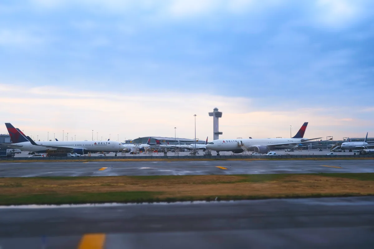 Two Delta Air Lines aircraft rest on the tarmac at John F. Kennedy International Airport as dusk settles over Queens. The iconic control tower stands sentinel in the background while runway lights begin to pierce the deepening twilight. Motion blur in the foreground suggests the constant movement of airport operations, while the serene blue-pink sky creates a striking contrast against the industrial aviation landscape.