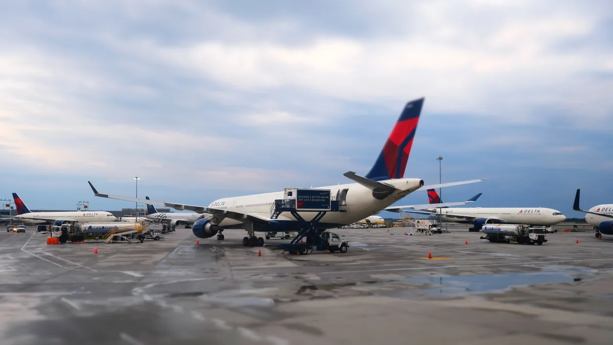 A Delta Air Lines wide-body aircraft dominates the rain-slicked tarmac at John F. Kennedy International Airport, its distinctive navy and red livery gleaming against the moody twilight sky. Ground support vehicles cluster around the aircraft while orange safety cones punctuate the wet concrete, creating a rhythmic pattern across the frame. The overcast heavens cast a silvery light that transforms the busy airport apron into an atmospheric tableau of modern aviation operations.