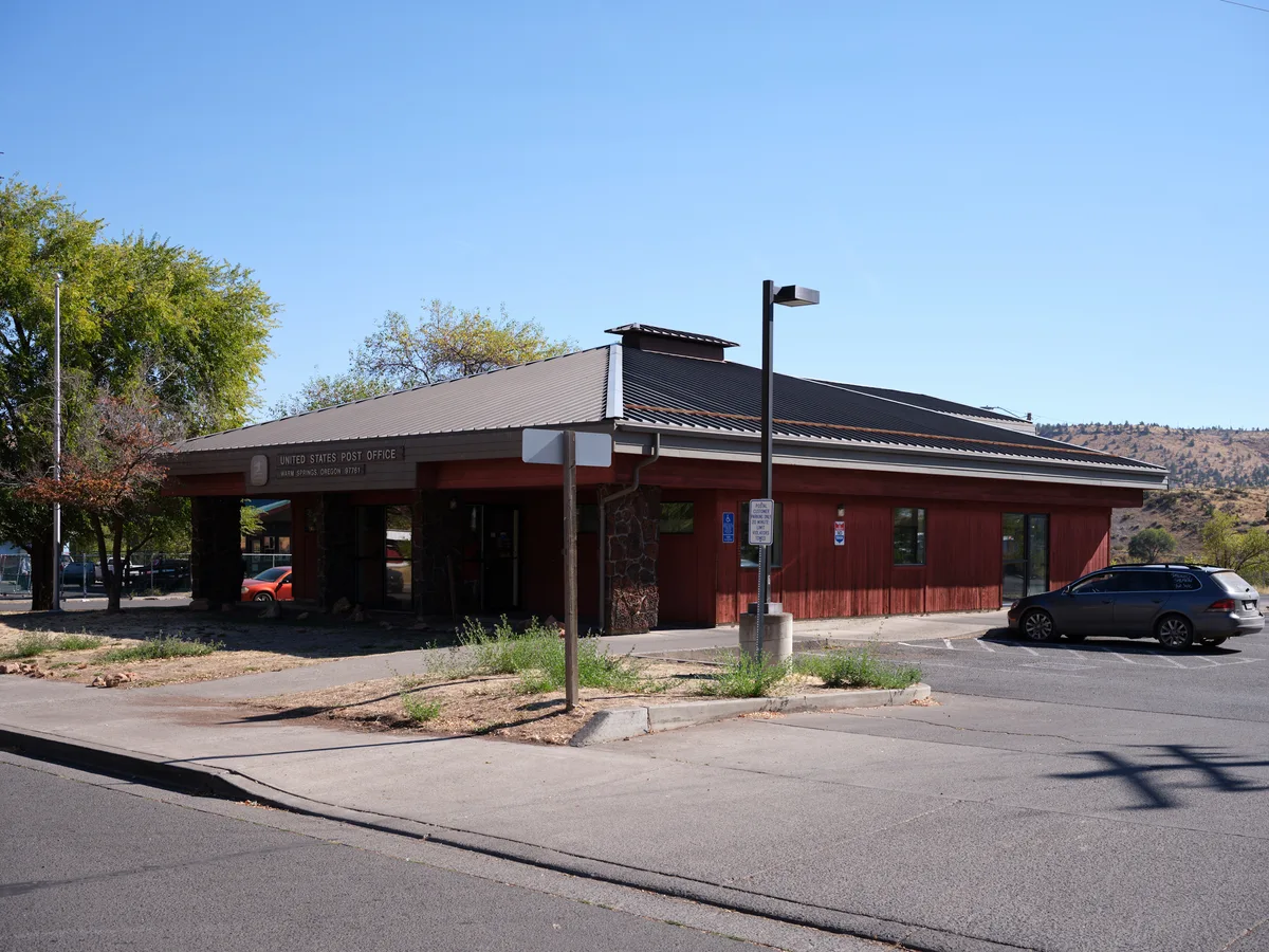 A weathered brick United States Post Office stands as a community anchor in Warm Springs, Oregon, its earth-toned facade harmonizing with the high desert landscape. The low-slung building features a distinctive overhanging roof and covered entrance, casting deep shadows across the front walkway under the brilliant blue sky. Scattered vehicles and sparse vegetation speak to the quiet rhythm of small-town life, while distant hills create a backdrop of muted autumn colors.