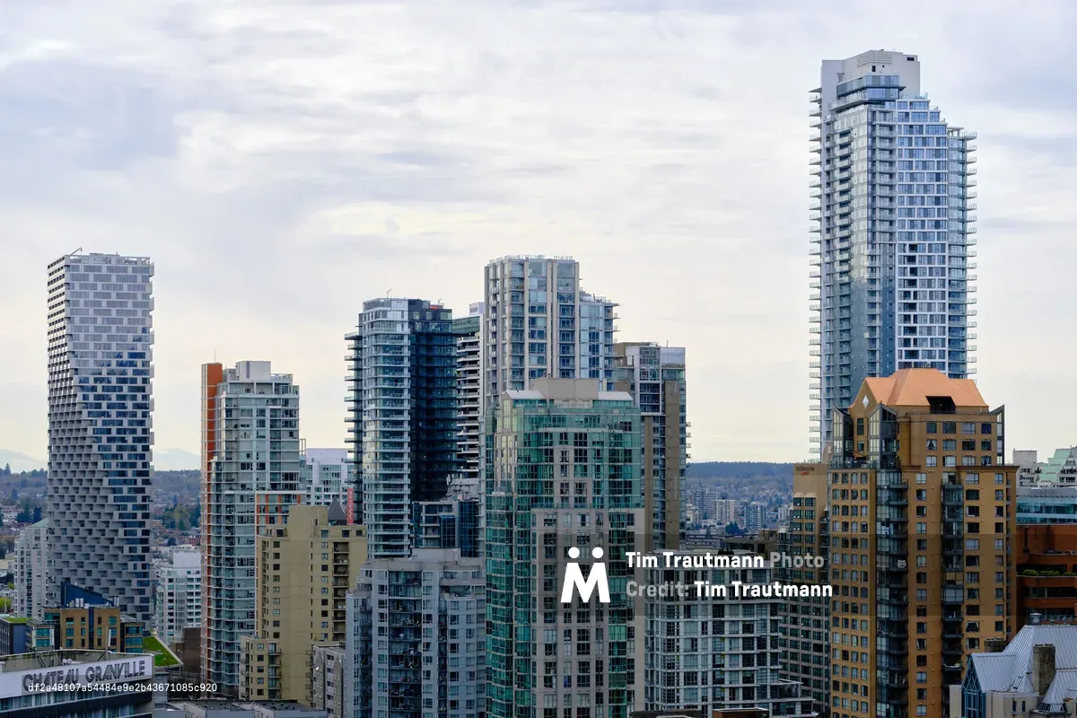 A dense cluster of contemporary residential towers rises against Vancouver's moody Pacific Northwest sky, their glass and concrete facades catching the diffused light of an overcast afternoon. The architectural composition showcases the city's modern urban density, with sleek high-rises in blues, grays, and warm terracotta tones creating a striking vertical landscape. In the foreground, the historic Chateau Granville sign anchors the scene, while forested hills stretch toward the horizon, emphasizing Vancouver's unique blend of urban sophistication and natural beauty.