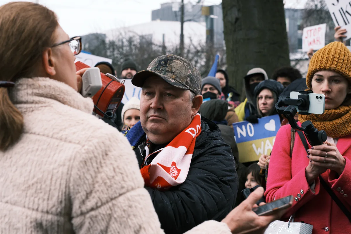 In the gray winter light outside Portland's Revolution Hall, tension crackles through a crowd gathered in the days before Russia's invasion of Ukraine. A middle-aged man in a camouflage cap stands resolute amid the pressing crowd, his weathered face betraying quiet determination as protesters surge around him with signs and smartphones raised. The intimate proximity of opposing voices creates a palpable friction, while bare trees and urban architecture frame this moment of civic discord on Southeast Stark Street.