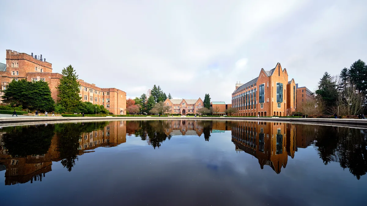 A perfectly still reflecting pool mirrors the iconic red brick architecture of the University of Washington campus under an overcast Pacific Northwest sky. The scene captures both historic collegiate Gothic buildings on the left and the modern angular forms of newer academic structures on the right, their warm terracotta facades creating a harmonious architectural dialogue. Towering evergreens frame the composition while scattered students traverse the distant pathways, adding human scale to this serene academic sanctuary. The glassy water surface doubles the visual impact, creating a symmetrical dreamscape where sky and earth converge.