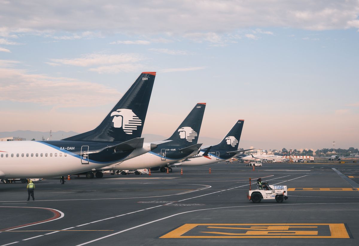 Three Aeroméxico passenger jets are parked in a row on the tarmac at Mexico City International Airport (Aeropuerto Internacional Benito Juárez), their dark navy tail fins bearing the airline's distinctive Aztec eagle logo receding in perspective from left to right. The aircraft registration numbers XA-DAH, XA-AMK, and XA-CCO are visible on the fuselages. A ground crew worker in a high-visibility vest walks near the closest aircraft, and a small white tug vehicle operates in the foreground. Orange traffic cones and yellow tarmac markings are visible on the dark pavement. Additional aircraft from other airlines are parked in the distance. The sky is pale blue with soft pink-tinted clouds, suggesting early morning or late afternoon light.