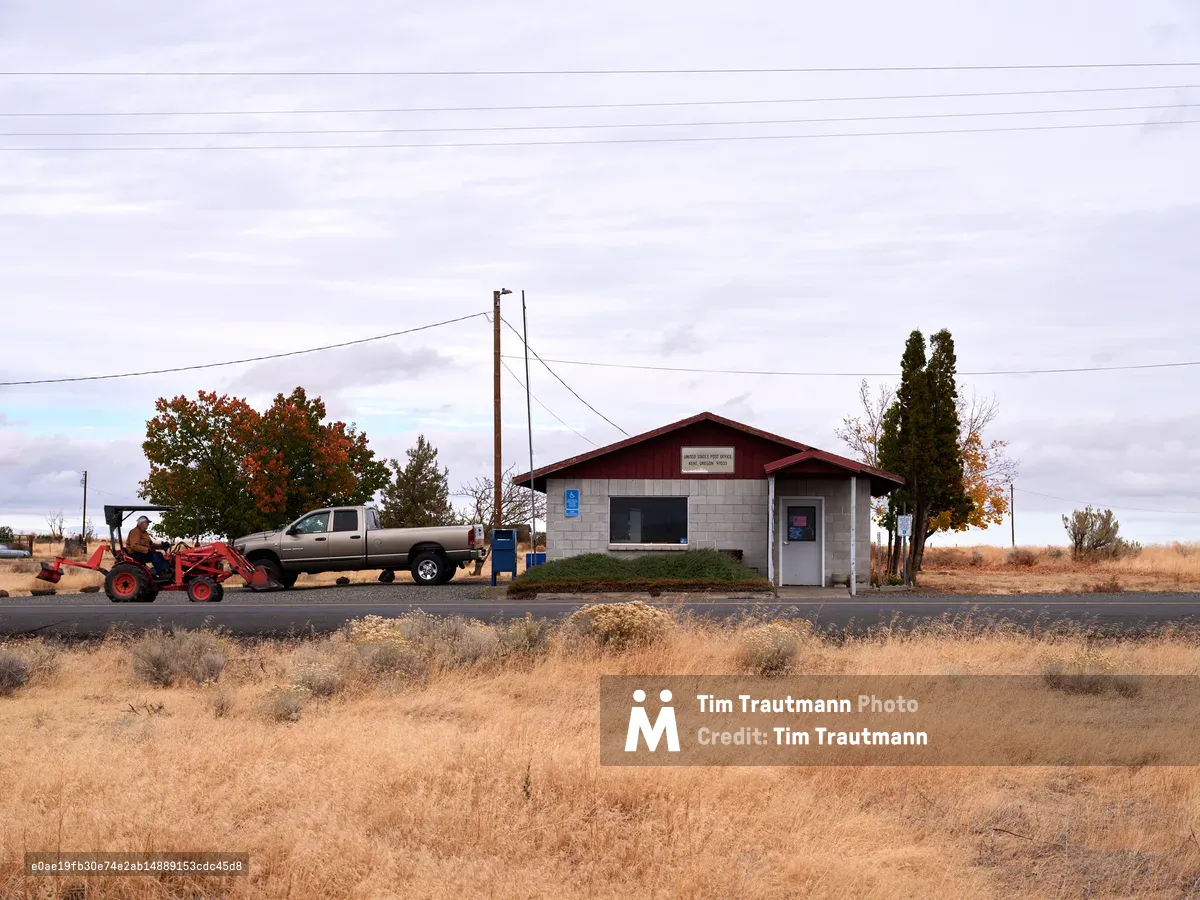 A modest United States Post Office sits beside a weathered asphalt road in Kent, Oregon, surrounded by the golden stubble of Sherman County's high desert landscape. The utilitarian building, with its burgundy metal roof and pale siding, serves as a humble anchor point in the vast expanse of eastern Oregon rangeland. A pickup truck with attached red tractor equipment idles nearby, while autumn-touched trees and utility lines punctuate the endless horizon under a brooding overcast sky.