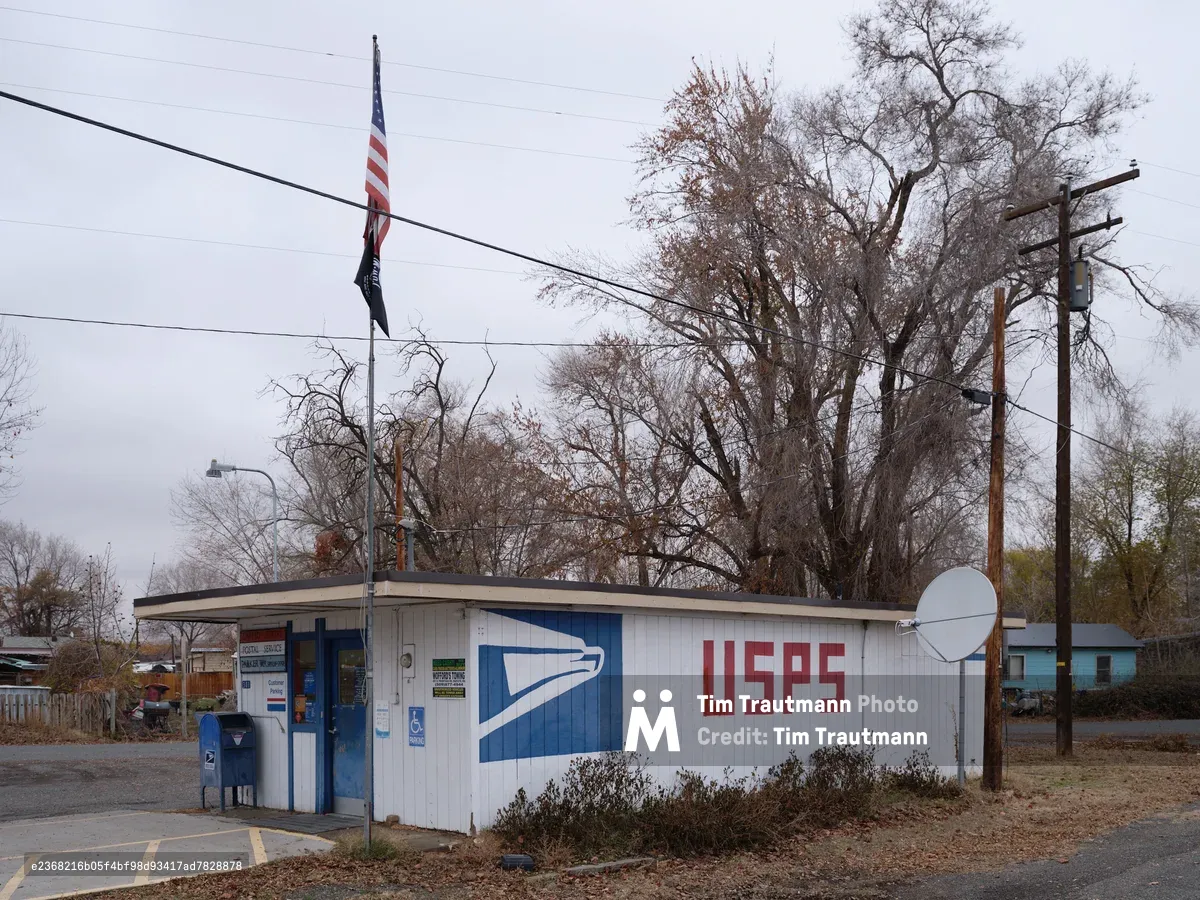A modest USPS facility stands beneath overcast winter skies in Parker, Washington, its white metal siding adorned with the familiar blue postal service logo and red lettering. Bare cottonwood trees loom behind the single-story building, their leafless branches creating a stark silhouette against the pewter sky. An American flag hangs motionless from a utility pole, while power lines crisscross the frame, emphasizing the rural isolation of this essential community service hub.