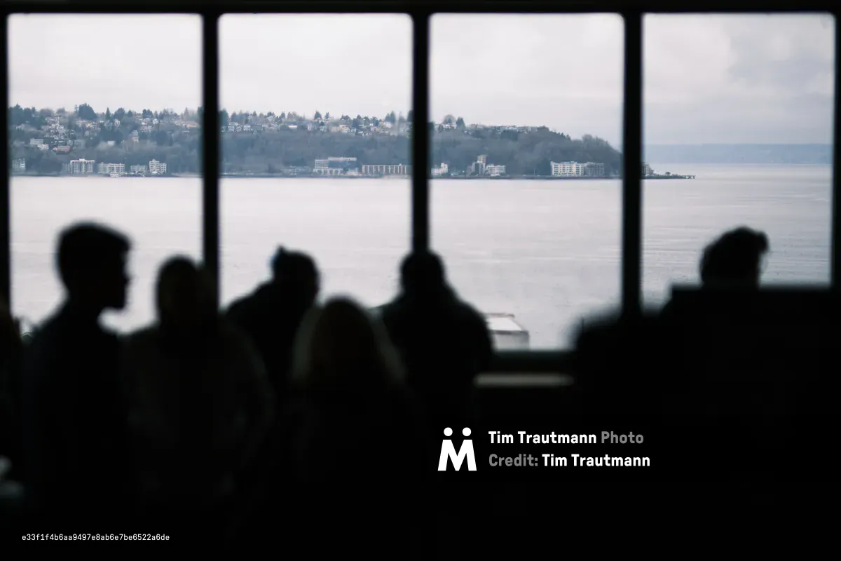 Silhouetted passengers aboard a Washington State Ferry peer through large windows toward the forested hillsides and residential developments of West Seattle across Elliott Bay. The atmospheric haze creates layers of depth, with modern apartment buildings and scattered homes climbing the verdant slopes. The composition frames human contemplation against the Pacific Northwest's signature blend of urban development and natural landscape, captured in soft, overcast light.