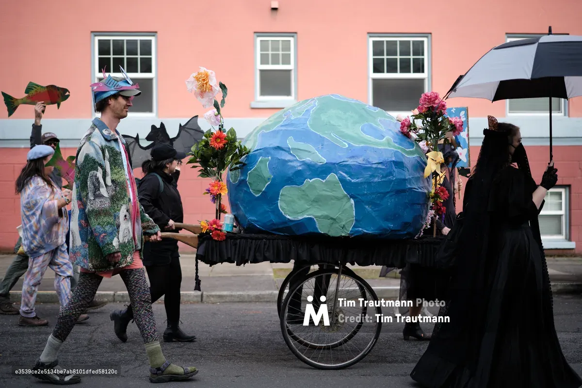 A group of costumed protesters participates in an environmental demonstration, wheeling a large papier-mâché model of Earth decorated with flowers on a cart through the streets of Portland, Oregon. Participants wear various costumes including nature-themed outfits and carry props while walking past coral-colored buildings.