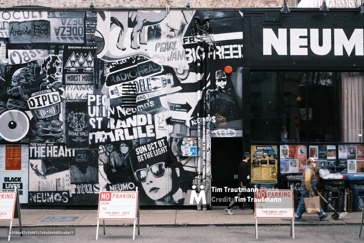 A weathered brick wall in Seattle's Capitol Hill neighborhood serves as a sprawling canvas for layered street art and concert posters, creating a dense collage of black, white, and red imagery. The anarchic composition includes band names like Modest Mouse and Neumos, political messages, and wheat-paste portraits that speak to the district's countercultural identity. Two pedestrians navigate the sidewalk past the artistic chaos, while red 'No Parking' signs punctuate the urban tableau with bureaucratic precision.