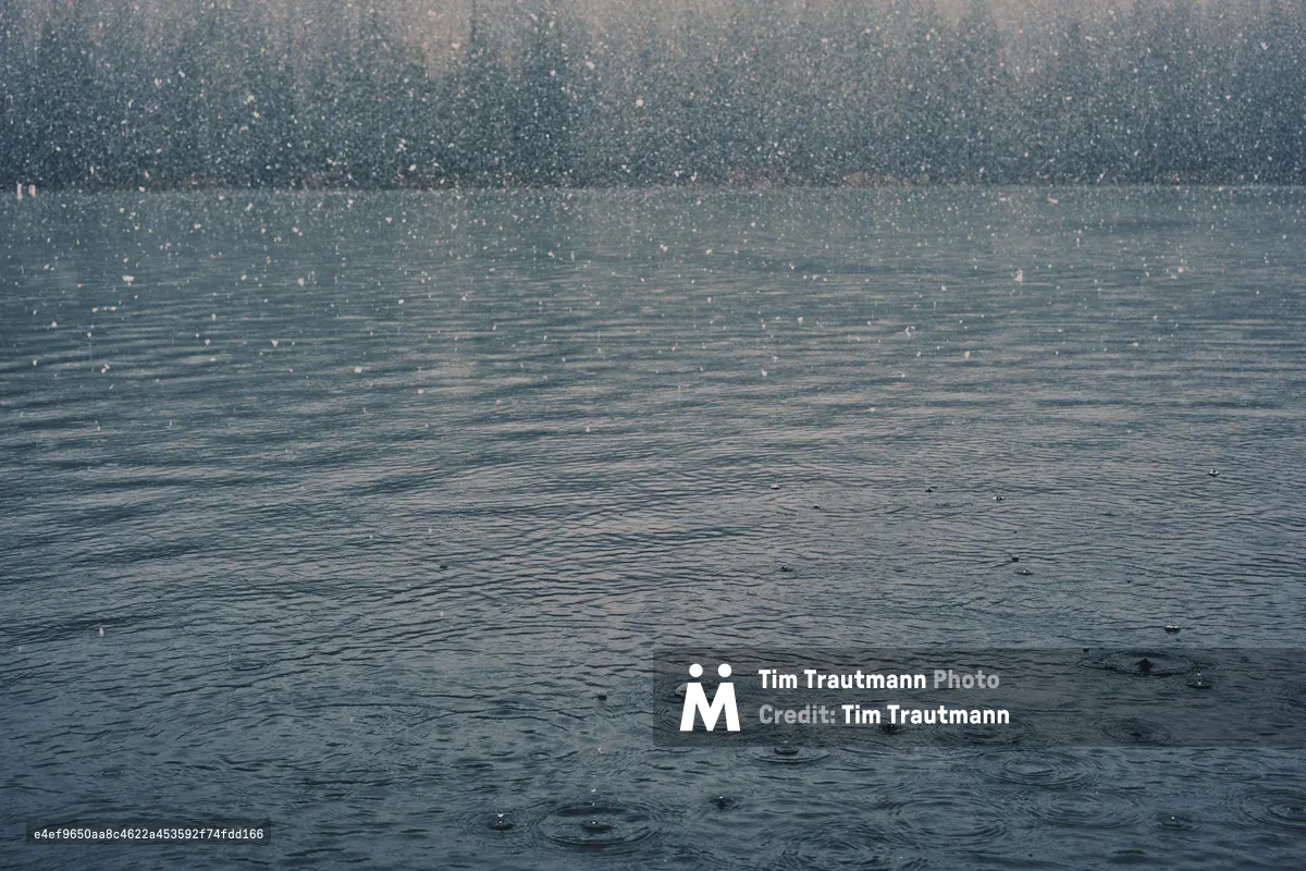 Countless raindrops create ephemeral ripples across the steel-gray surface of a Central Oregon lake near Sisters. The atmospheric precipitation transforms the water into a textured canvas of concentric circles, each drop's impact sending delicate rings outward before disappearing into the moody, overcast scene. The misty air above the lake blurs with falling rain, creating a dreamlike quality that speaks to the Pacific Northwest's contemplative weather patterns.