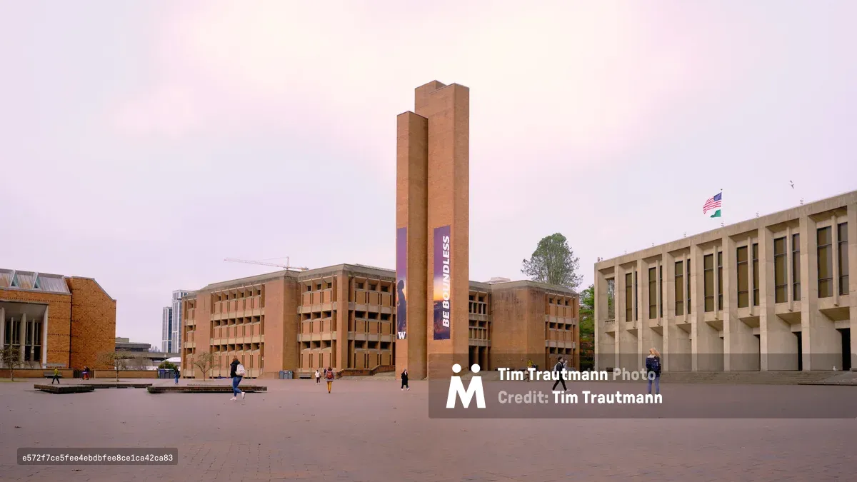 The iconic Red Square at the University of Washington bathes in ethereal pink twilight, with Louis Kahn's brutalist Suzzallo Library tower standing sentinel against a pastel sky. Students traverse the vast brick plaza in scattered formation, their figures dwarfed by the monumental concrete and brick architecture that defines this celebrated campus heart. The geometric interplay between the library's vertical thrust and the horizontal expanse of surrounding academic buildings creates a striking study in modernist design, while the soft evening light transforms the utilitarian materials into something almost mystical.