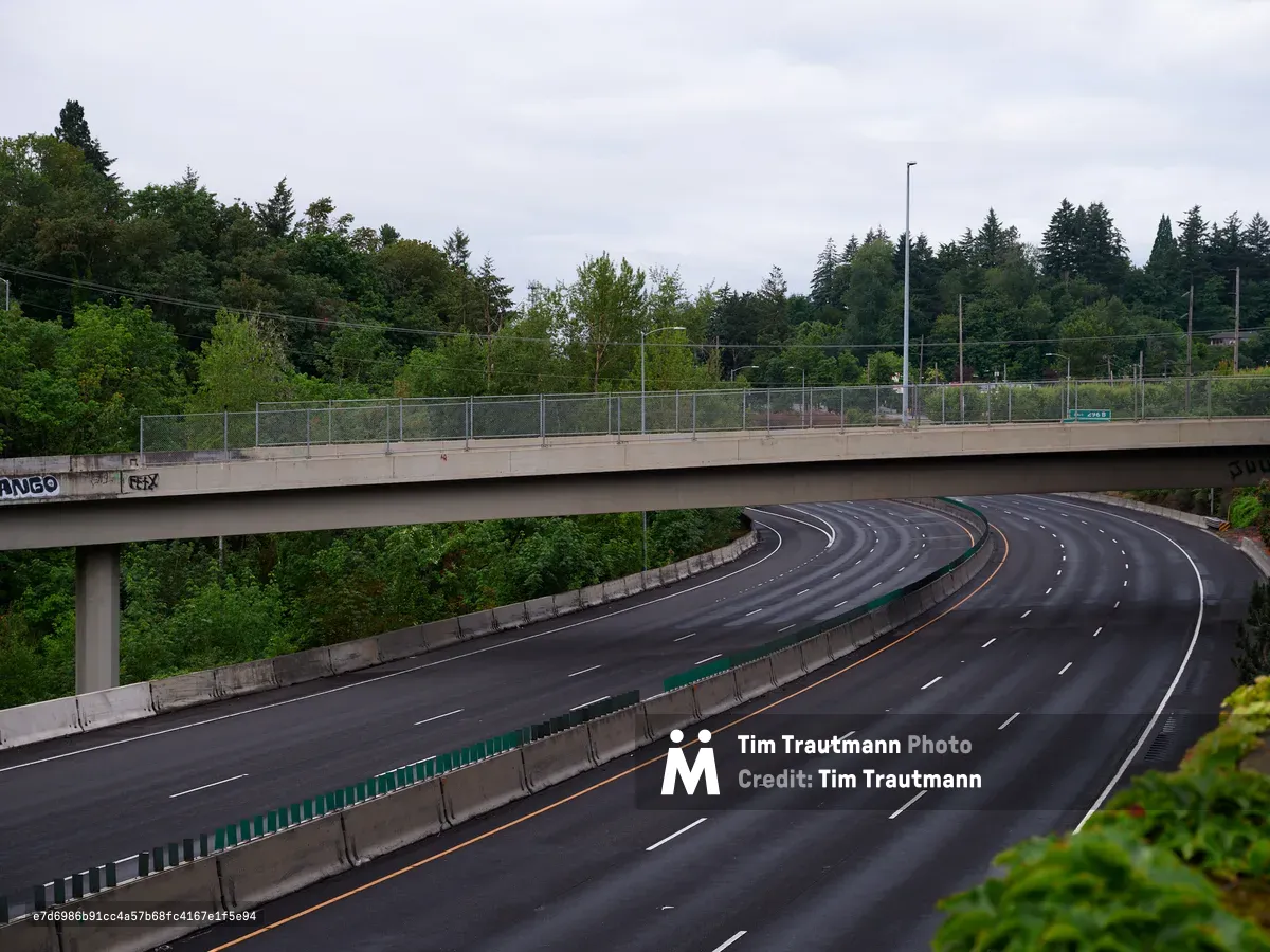A curved section of multi-lane highway passes under a concrete pedestrian overpass surrounded by lush Pacific Northwest forest. The wet asphalt road features white lane markings and orange striping along the median barrier, with dense green trees creating a natural canopy overhead under an overcast sky.