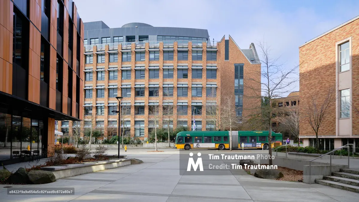 A King County Metro articulated bus glides through the University of Washington campus plaza, its green and gold livery creating a striking counterpoint to the warm brick facades of modern academic buildings. The late afternoon light casts gentle shadows across the concrete courtyard, where bare winter trees stand sentinel among carefully planned landscaping. The architectural composition frames the scene with geometric precision, balancing the institutional grandeur of the multi-story research buildings against the human scale of the transit corridor.