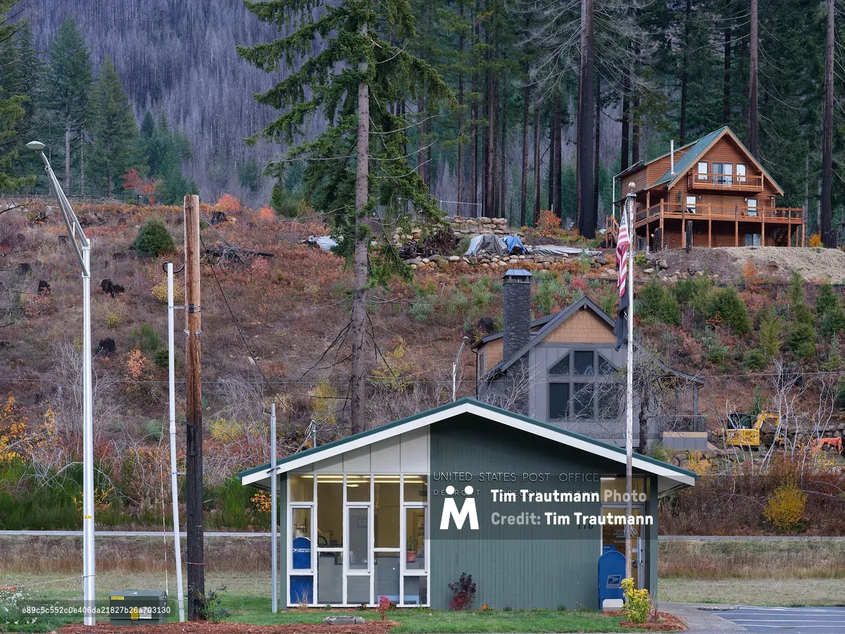 A small green and white single-story United States Post Office building in Detroit, Oregon 97342, with an American flag on a pole out front and a blue USPS mailbox beside the entrance. Behind it, a hillside shows dramatic evidence of wildfire damage — charred tree stumps, scorched earth, and burnt vegetation in rusty oranges and browns. A surviving log cabin-style home with a green roof sits among tall evergreen pines on the hillside above. Construction equipment is visible to the right, suggesting ongoing recovery work. The moody, overcast scene conveys a community in the process of rebuilding.
