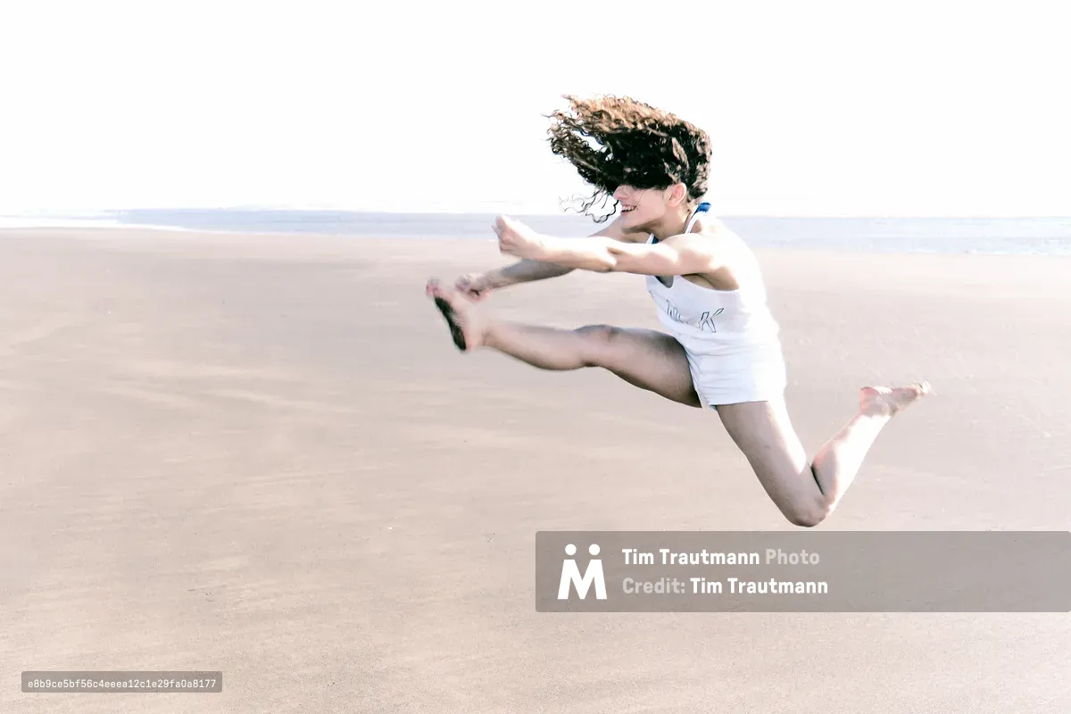 A young woman leaps joyfully into the air on a wide, flat sandy beach, her curly dark hair flying across her face. She wears a white tank top and shorts, arms outstretched and legs bent mid-jump, laughing as she soars above the sand. The bright, overcast sky and pale beach stretch to the horizon behind her.