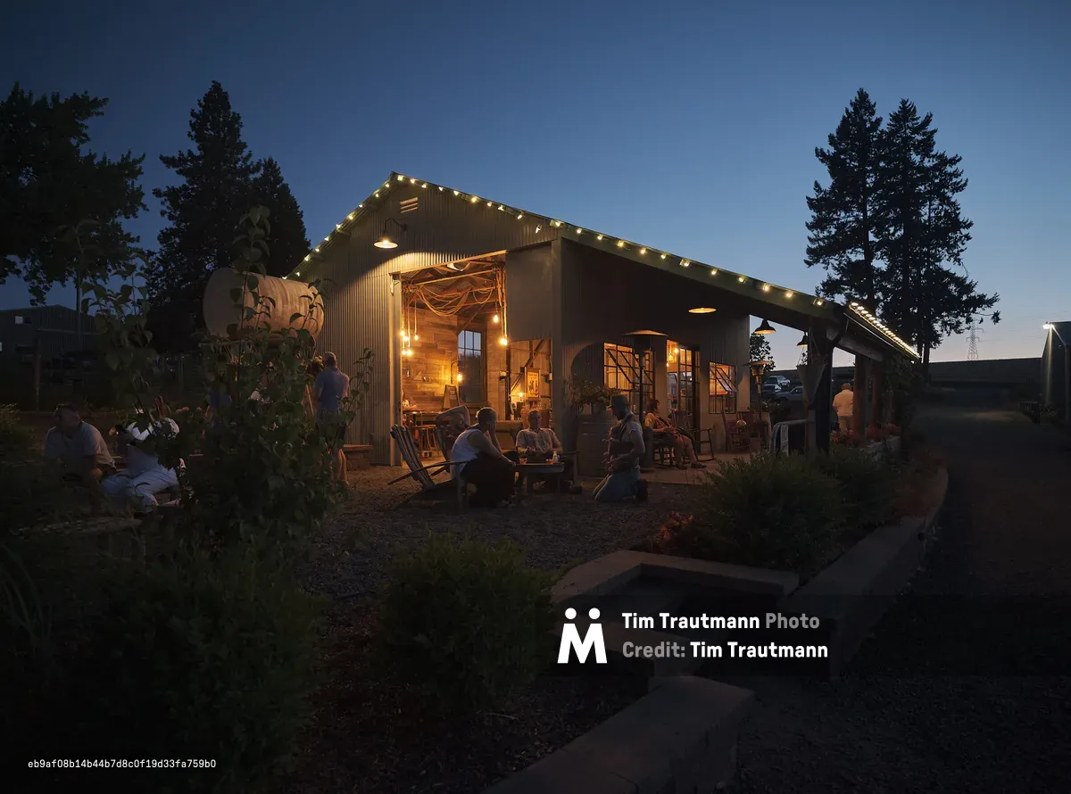 The exterior of the barn at Abbey Road Farm in Carlton, Oregon, photographed at dusk. The corrugated metal barn is strung with warm globe lights along its roofline, glowing invitingly against a deep blue twilight sky. Groups of guests relax on Adirondack chairs and benches on the gravel patio, socializing in the warm light spilling from the open barn interior. Tall evergreen trees flank the building, and lush garden plantings border a stone pathway in the foreground.