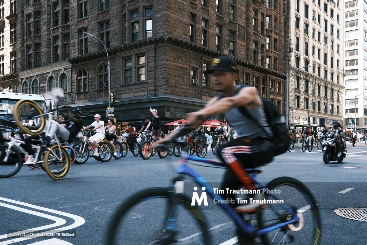 Motion blur captures the kinetic energy of dozens of cyclists flowing down 7th Avenue in Midtown Manhattan, their forms ghosted against the solid backdrop of historic brownstone architecture. The foreground rider in a blue cap emerges sharp from the stream of blurred movement, while golden-rimmed bicycle wheels catch fragments of afternoon light. Behind the cycling parade, the stately Carnegie Hall neighborhood's characteristic sandstone facades provide timeless contrast to this modern urban ballet of sustainable transportation.