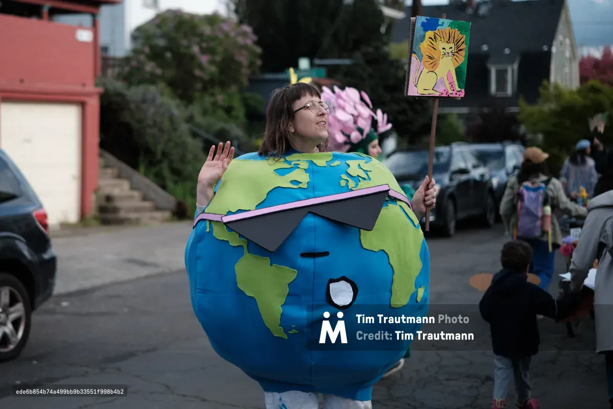 A person wearing a large spherical Earth costume with blue oceans and green continents stands on a Portland street, holding a protest sign while participating in what appears to be a climate change demonstration. The costume features sunglasses and the person is waving at the camera.