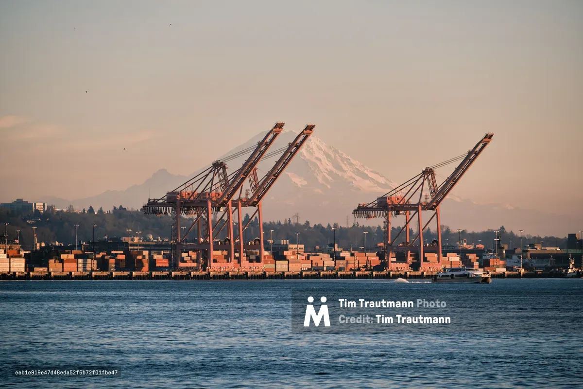 The majestic snow-capped peak of Mount Rainier dominates the horizon behind Seattle's bustling container port, where towering orange gantry cranes stand sentinel over geometric stacks of cargo containers. Golden hour light bathes the industrial waterfront in warm amber tones, while gentle ripples disturb Elliott Bay's surface as a small vessel navigates the foreground. The composition creates a striking juxtaposition between Washington's natural grandeur and the relentless pulse of maritime commerce.