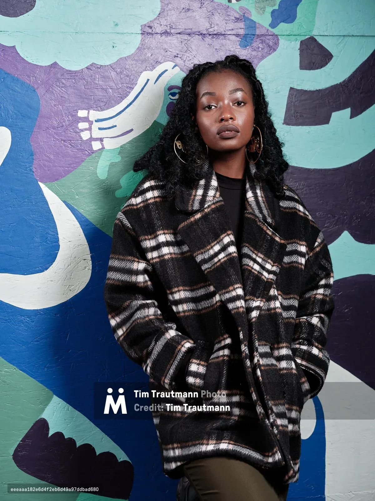 A portrait of artist Naomi Likayi standing against a vibrant painted mural in teal, blue, purple, and white. She wears an oversized black and white plaid wool coat over a black top, with olive trousers and large gold hoop earrings. Her long black braids frame her face as she gazes directly into the camera with a calm, composed expression.