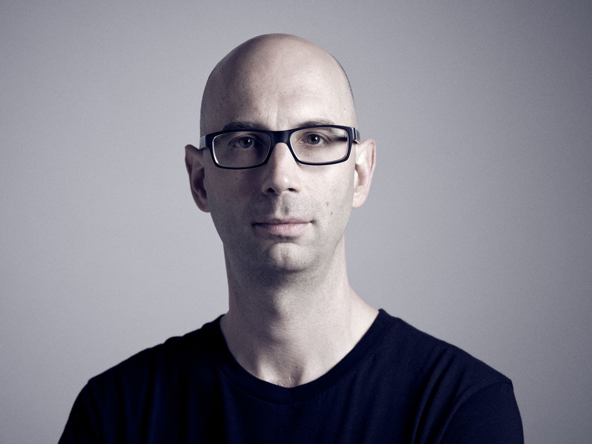 DJ and electronic music producer Jack Coleman sits for an intimate studio portrait against a refined gray backdrop in Portland's creative Central Eastside district. The controlled lighting creates subtle shadows that sculpt his bald head and black-framed glasses, while his dark v-neck shirt dissolves into shadow, focusing attention on his contemplative expression. The minimalist composition reflects the precision and artistry of his electronic music work with labels like Trapez and Translucent.