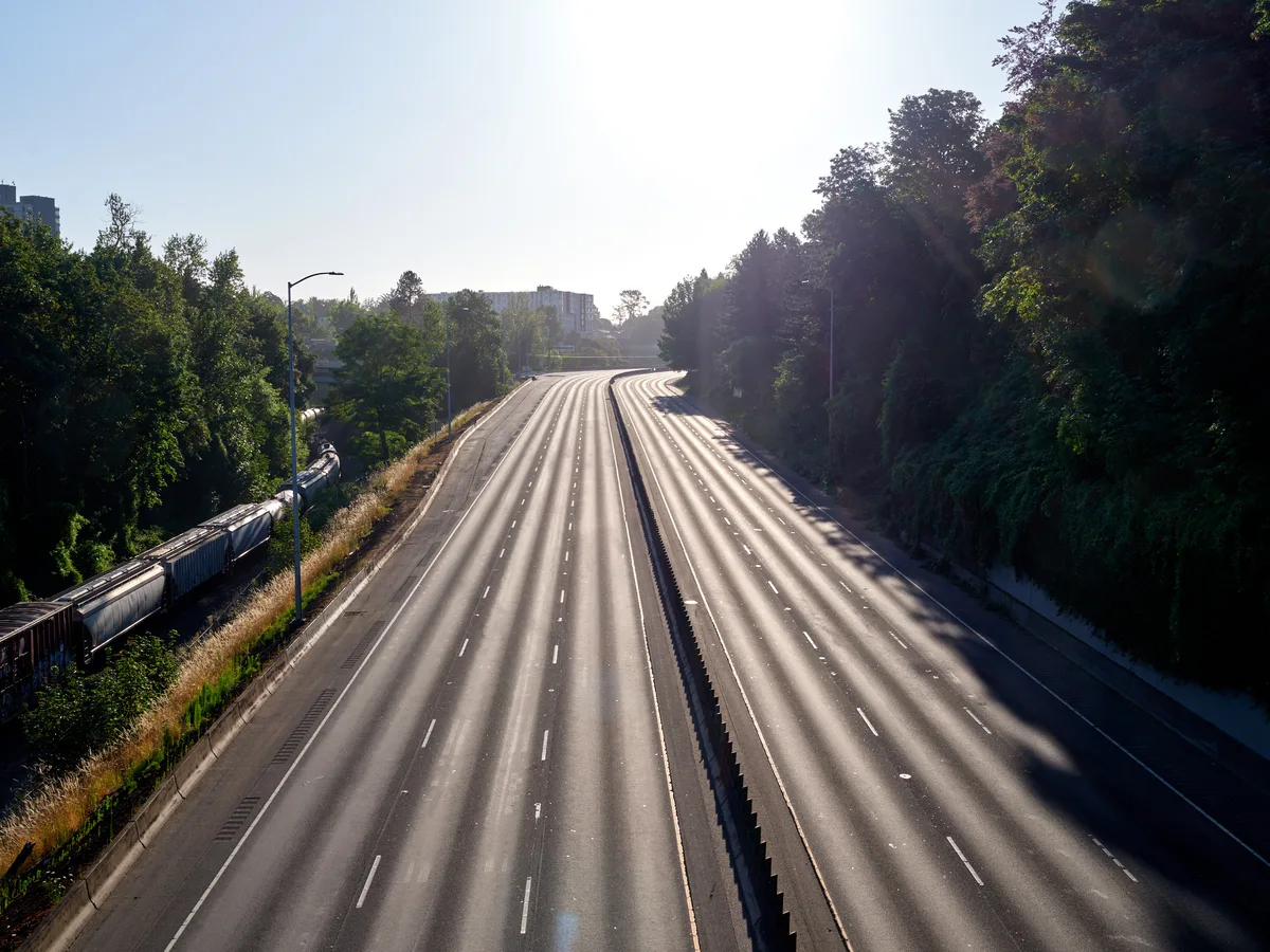 An aerial view of the empty Banfield Freeway (Interstate 84) in Portland's Lloyd District, showing multiple lanes of asphalt stretching into the distance with lush green trees lining both sides and urban buildings visible in the background under bright sunlight.