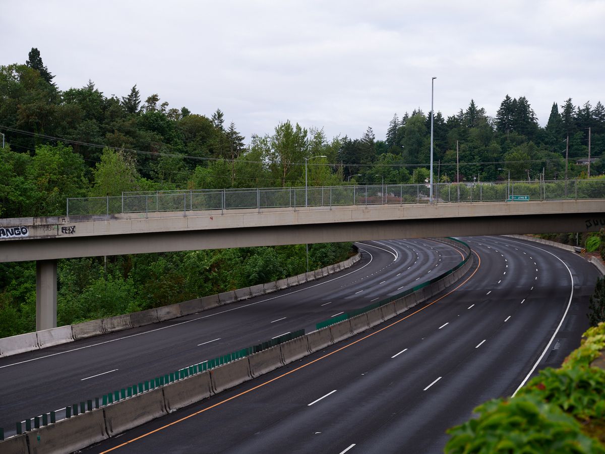 A curved section of multi-lane highway passes under a concrete pedestrian overpass surrounded by lush Pacific Northwest forest. The wet asphalt road features white lane markings and orange striping along the median barrier, with dense green trees creating a natural canopy overhead under an overcast sky.