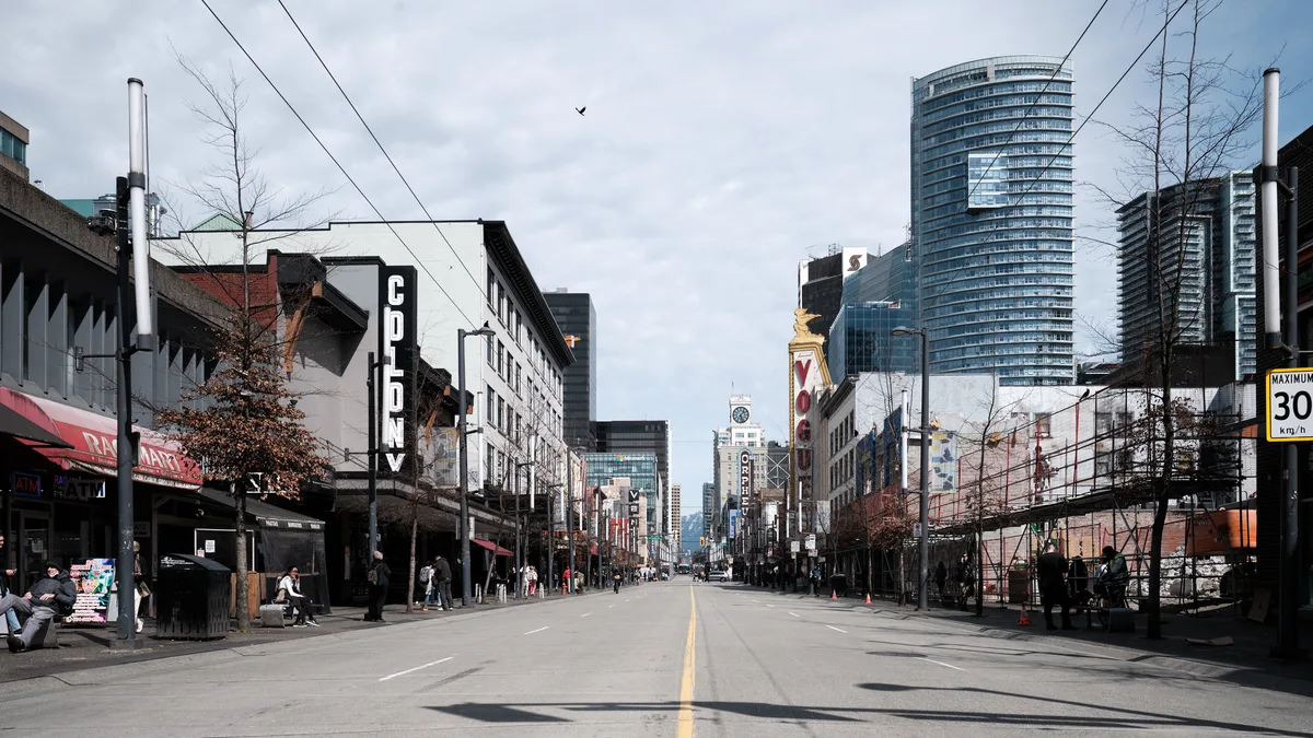 A deserted asphalt boulevard stretches toward Vancouver's downtown core, flanked by the eclectic mix of heritage storefronts and soaring glass towers that define the city's evolving skyline. The Colony Theatre's bold vertical signage punctuates the left side of the frame, while modern residential high-rises pierce the overcast sky in the distance. Sparse pedestrians and the conspicuous absence of traffic create an unusual quietude along this typically bustling commercial artery, with power lines creating geometric patterns overhead against the muted gray clouds.