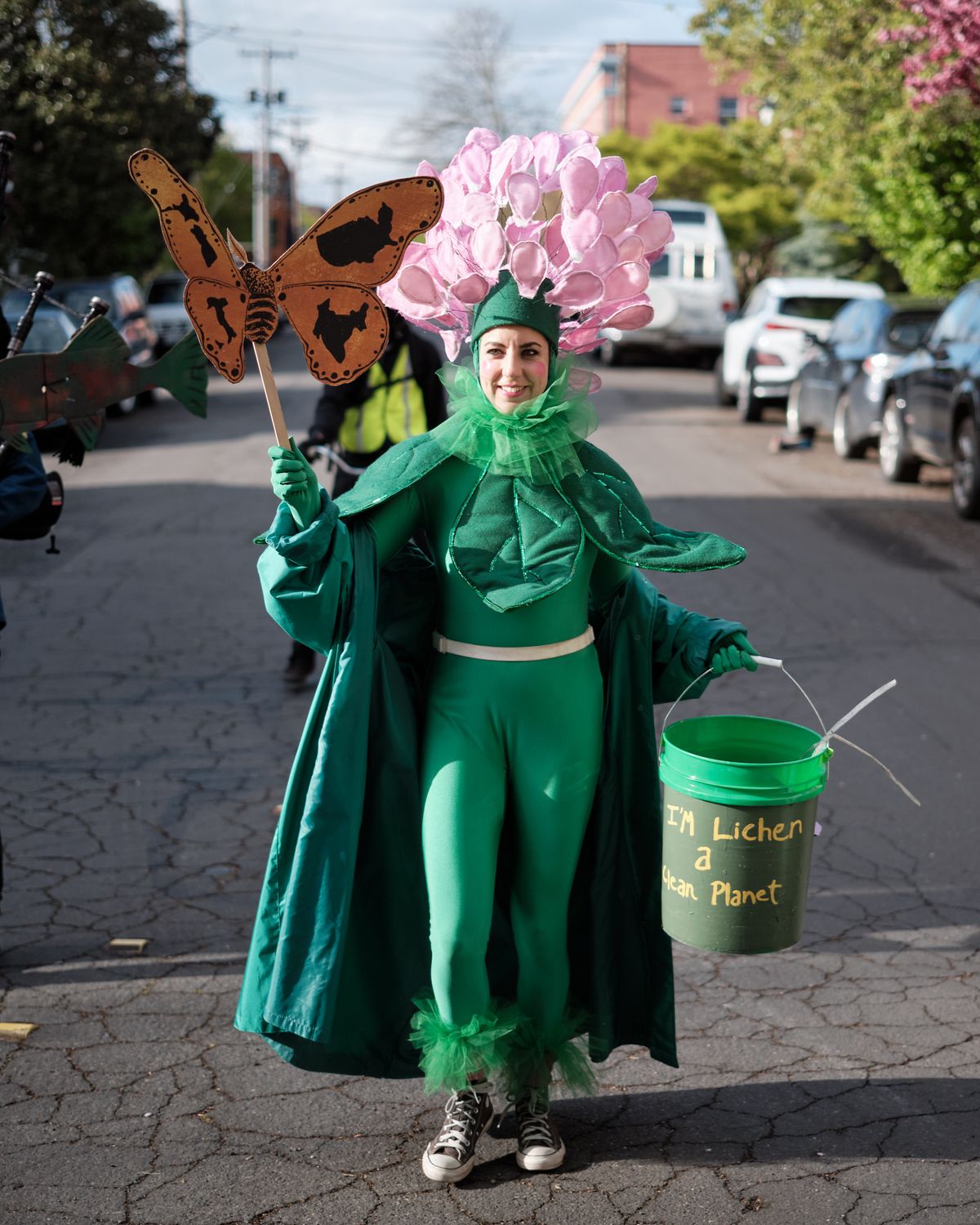 A person dressed in an elaborate green plant costume with pink flowers stands on a cobblestone street in Portland, Oregon, holding a butterfly prop and a bucket with environmental messaging. The costume includes leafy details and the bucket reads about creating a clean planet.