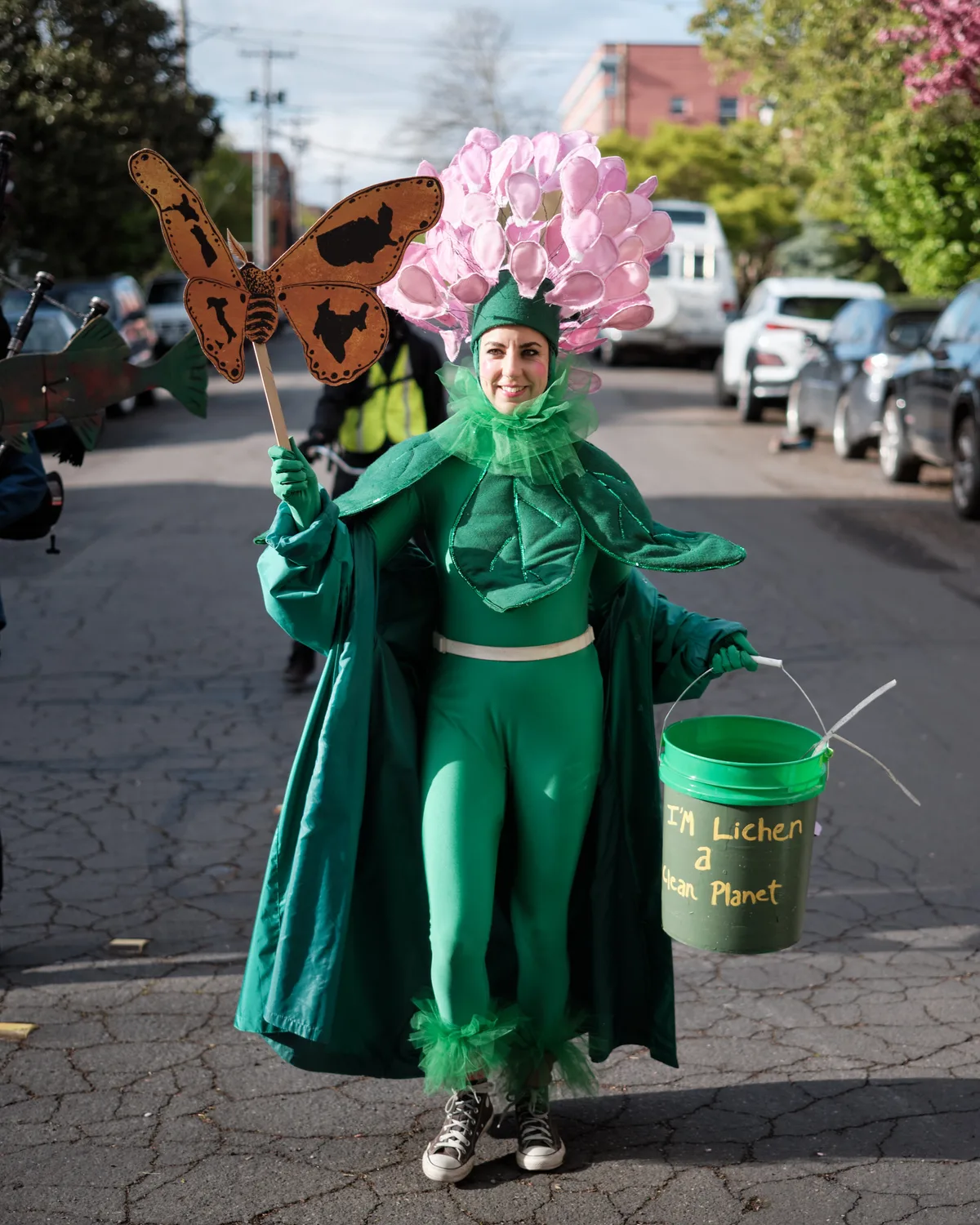 A person dressed in an elaborate green plant costume with pink flowers stands on a cobblestone street in Portland, Oregon, holding a butterfly prop and a bucket with environmental messaging. The costume includes leafy details and the bucket reads about creating a clean planet.