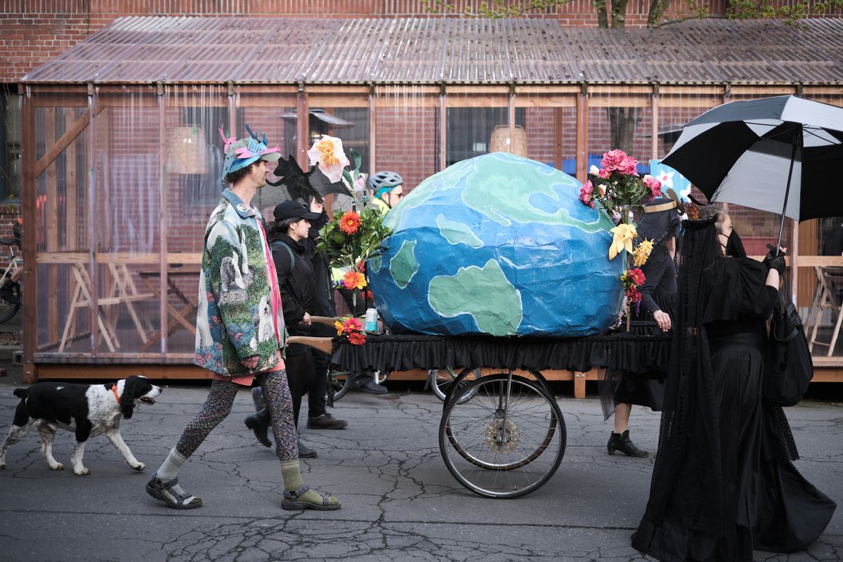A group of mourners dressed in black carry a large blue and green Earth-shaped coffin on a wheeled cart through the streets of Portland, Oregon, accompanied by flowers and an umbrella in what appears to be an environmental protest or artistic funeral procession.