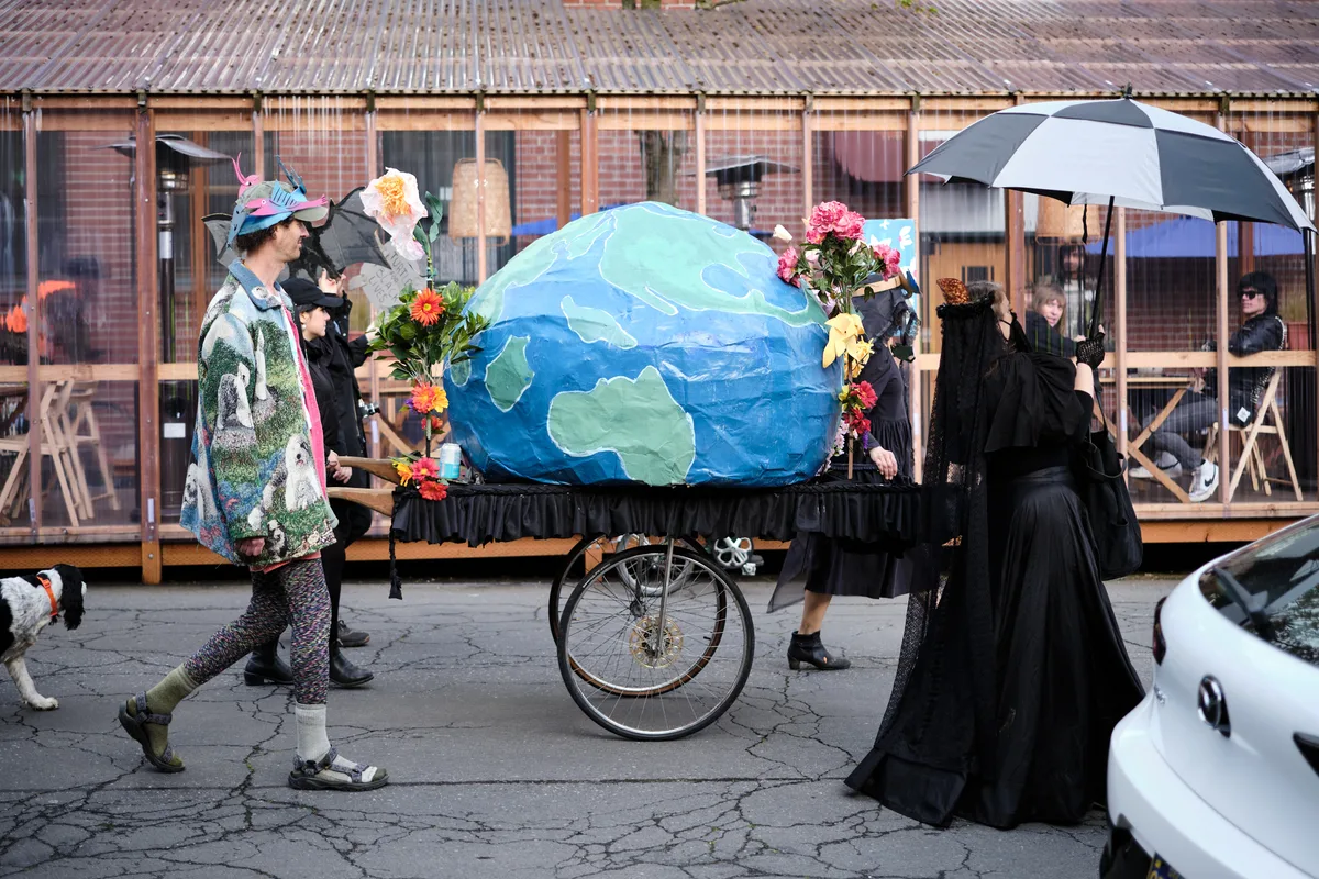 A group of people in mourning attire participate in an environmental protest or performance art piece, wheeling a large painted Earth globe on a funeral cart through the streets of Portland's Central Eastside district. The participants wear black clothing and carry flowers, creating a somber funeral procession for the planet.