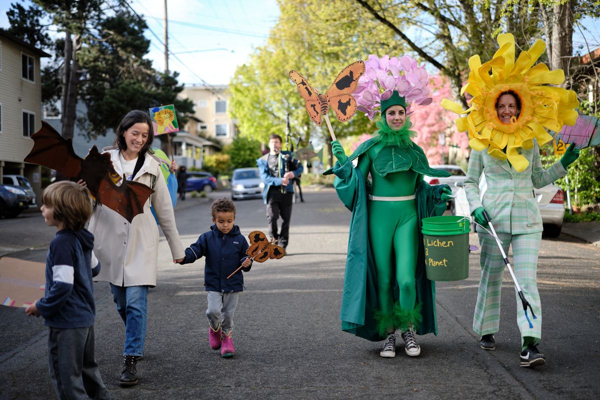 A group of environmental activists and families participate in a colorful street parade in Portland, Oregon, featuring participants dressed as flowers, trees, and nature-themed costumes while carrying eco-friendly signs and props. Children and adults walk together on a residential street lined with trees and houses, promoting environmental awareness through creative costumes and community engagement.