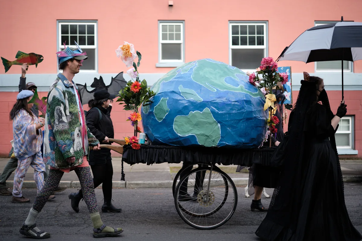 A group of costumed protesters participates in an environmental demonstration, wheeling a large papier-mâché model of Earth decorated with flowers on a cart through the streets of Portland, Oregon. Participants wear various costumes including nature-themed outfits and carry props while walking past coral-colored buildings.