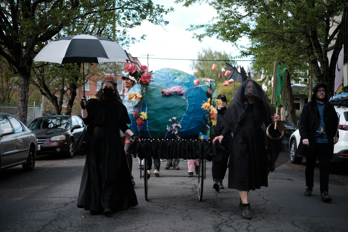 A group of protesters dressed in black robes march down a tree-lined street in Portland's Central Eastside district, carrying a large blue Earth globe decorated with flowers on a wheeled cart. The solemn procession appears to be an environmental demonstration or funeral-themed climate protest, with participants holding umbrellas and wearing dark clothing while walking among parked cars on Southeast 14th Avenue.