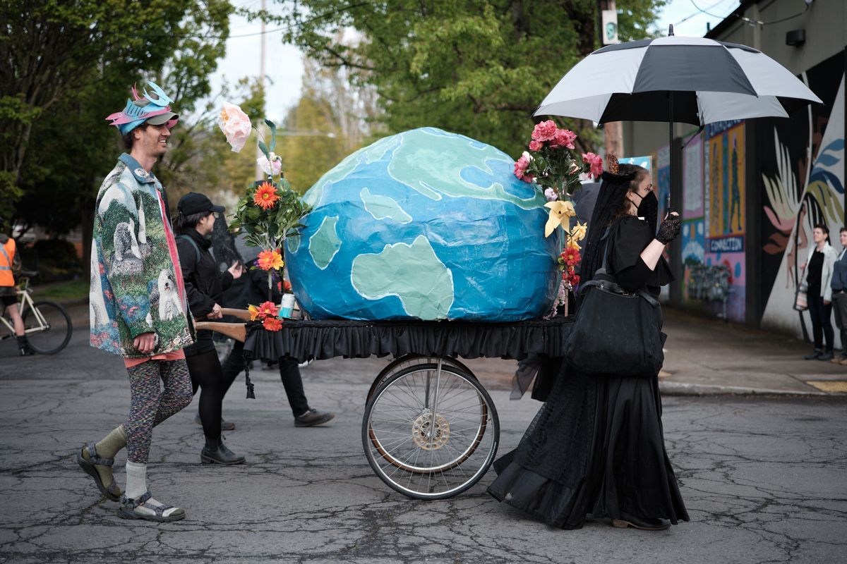 A group of environmental activists in Portland, Oregon participate in a street demonstration, pushing a wheeled cart carrying a large handmade model of Earth decorated with flowers. The protesters are dressed in colorful, eclectic clothing and one holds an umbrella, suggesting a community-organized environmental awareness event.