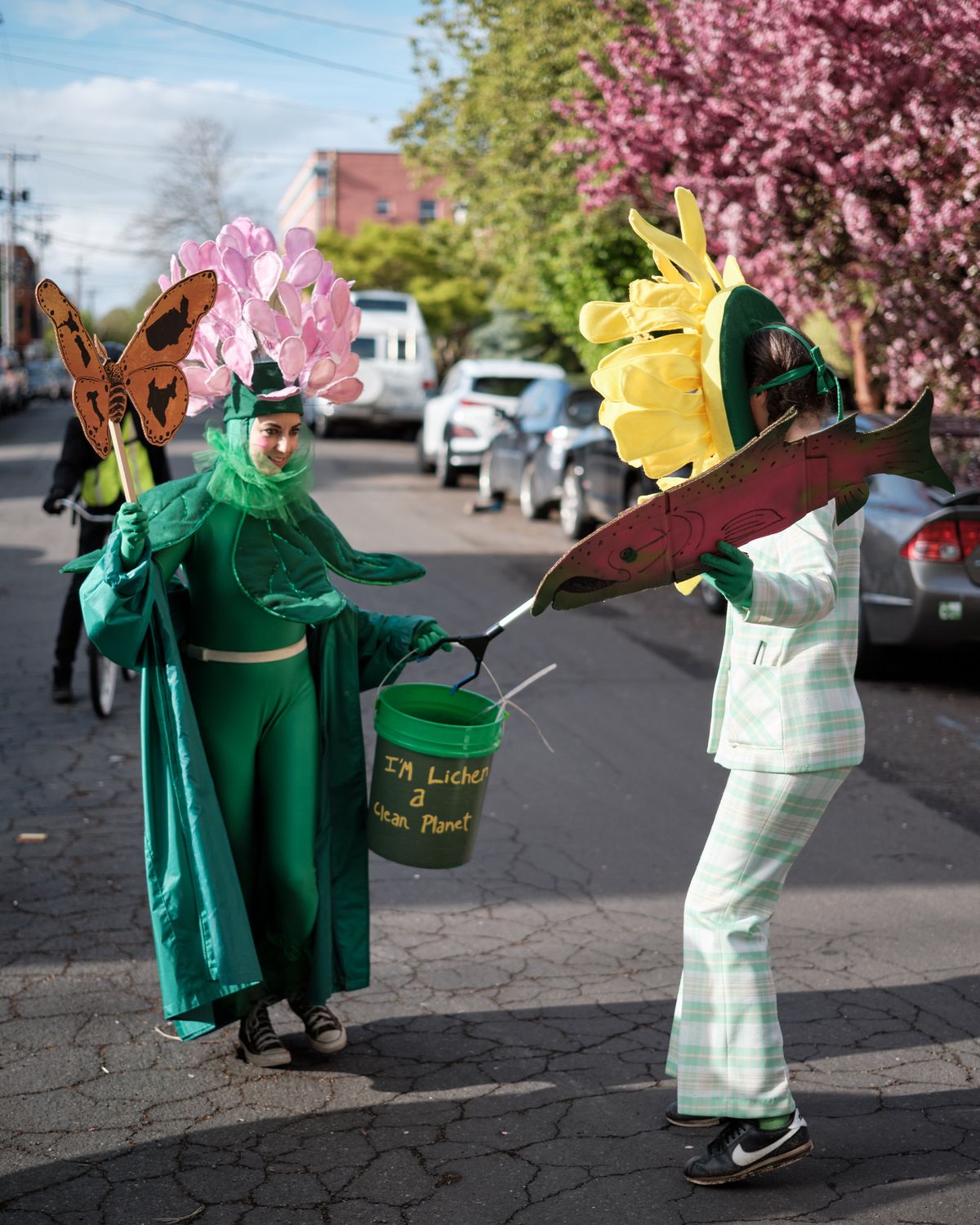 Two costumed street performers in Portland, Oregon engage in an environmental awareness demonstration, with one person dressed as a flower holding a bucket labeled 'I'm Lichen a Clean Planet' and another wearing nature-themed props including butterfly and fish cutouts.