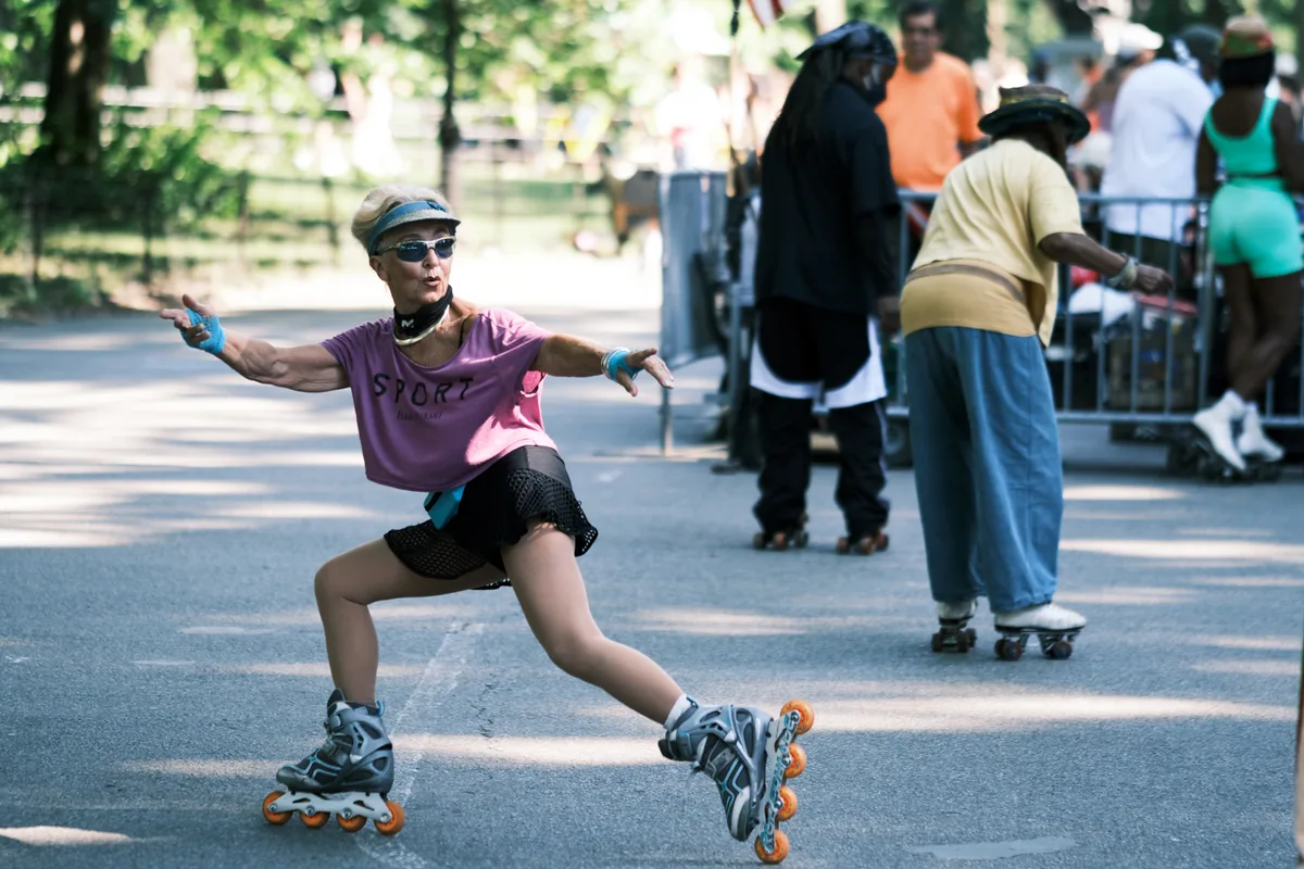 A joyful woman in purple athletic wear and sunglasses glides through Central Park on inline skates, her arms spread wide in triumphant celebration. Dappled sunlight filters through the leafy canopy above, creating a warm, golden atmosphere on the park's paved pathways. Behind her, other park visitors stroll leisurely, creating a perfect contrast between her dynamic energy and the peaceful weekend ambiance. The composition captures the pure freedom and exhilaration of urban recreation in Manhattan's beloved green oasis.