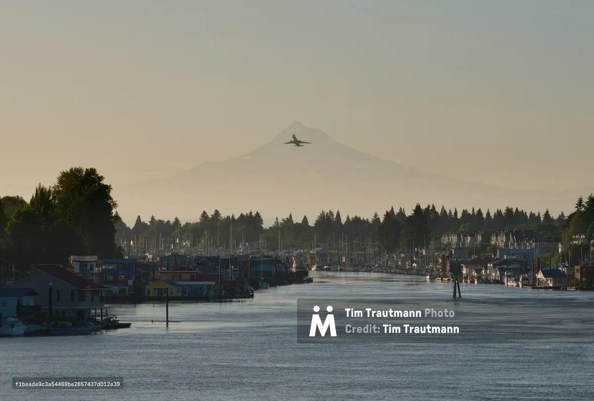 A commercial aircraft cuts through the pale morning sky above Mount Hood's ghostly silhouette, while the Columbia River winds past Portland's North Harbor in the golden hour light. The waterway reflects the muted tones of dawn as fishing boats and houseboats line the quiet marina, their masts creating a delicate forest against the distant Cascade Range. This serene moment captures the intersection of urban aviation and maritime life, where the rhythms of river commerce play out beneath the watchful presence of Oregon's iconic peak.
