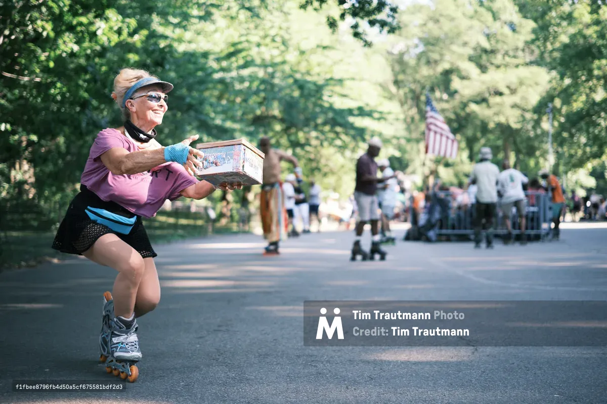 A spirited woman in a pink top and black athletic wear gracefully rollerskates along Central Park's tree-lined pathways, carrying what appears to be a lunch cooler. Dappled sunlight filters through the dense canopy above, casting dancing shadows on the asphalt as she navigates past blurred pedestrians and a distant American flag. Her confident posture and protective gear speak to both skill and safety consciousness in this quintessential New York moment.