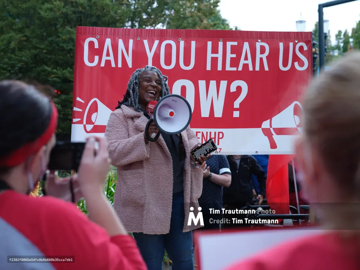 A determined healthcare worker with dreadlocks commands attention through a red and white megaphone, her infectious smile contrasting with the serious nature of the Kaiser Permanente nurses' strike in Portland's Lloyd District. Behind her, a bold crimson banner emblazoned with "CAN YOU HEAR US NOW?" amplifies her message to the sea of red-clad supporters gathered in the foreground. The late afternoon light filters through verdant trees, casting a warm glow over this moment of collective action outside the towering Kaiser Permanente headquarters on Northeast Multnomah Street.