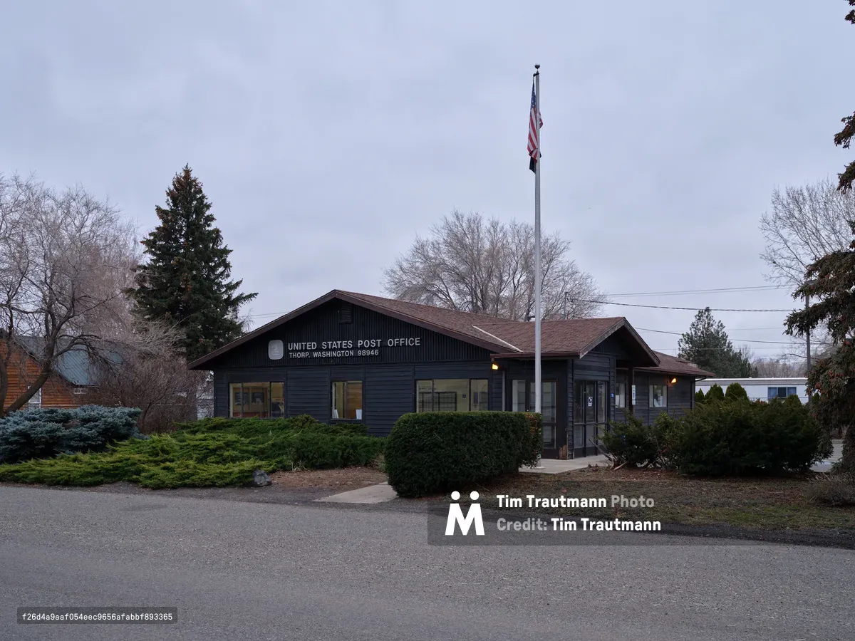 The modest United States Post Office in Thorp, Washington sits quietly beneath a pewter evening sky, its dark wood siding and warm interior glow creating an intimate portrait of small-town America. The single-story building, flanked by evergreen shrubs and bare winter trees, displays the Stars and Stripes on a tall flagpole that pierces the overcast horizon. Golden light spills from the windows onto the empty parking lot, suggesting the quiet continuity of postal service in this rural Kittitas County community.