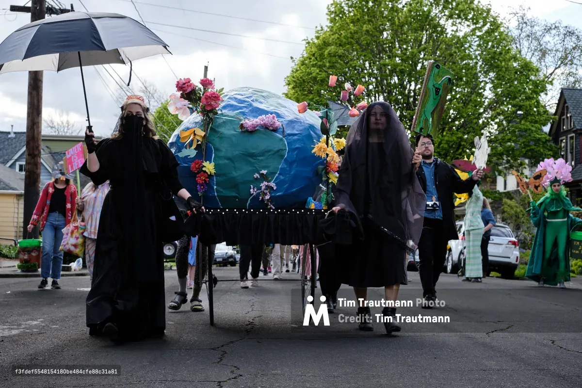 A group of environmental activists dressed in black participate in a community parade in Portland, Oregon, carrying a large Earth globe decorated with flowers on a wheeled cart while walking down a residential street lined with trees and houses.