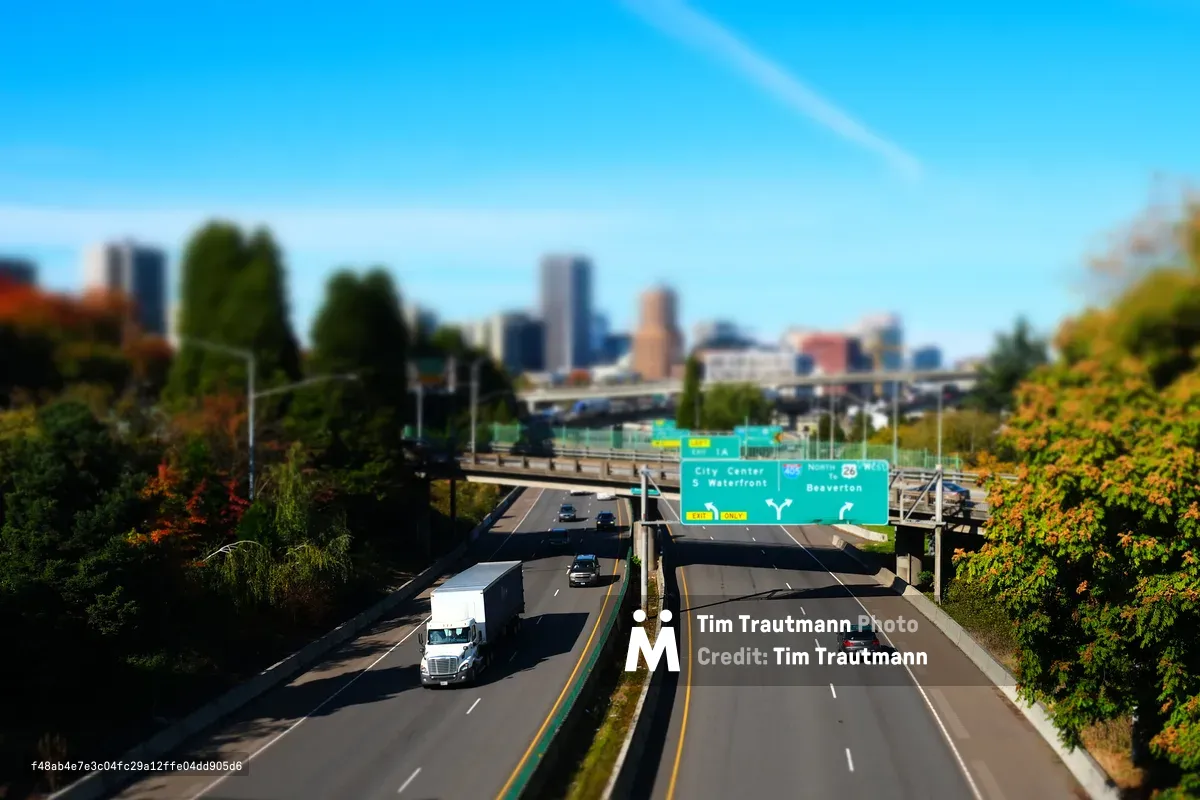 A tilt-shift photograph of Interstate 5 in South Portland, giving the scene a miniature toy-like quality. A white semi-truck and several cars travel northbound toward downtown Portland, whose skyline rises in the soft-focus background. Green highway signs direct traffic toward City Center, South Waterfront, and Beaverton via Route 26. Autumn foliage in orange, red, and green lines both sides of the freeway, and an overpass bridge and interchange infrastructure are visible in the middle distance under a vivid blue sky.
