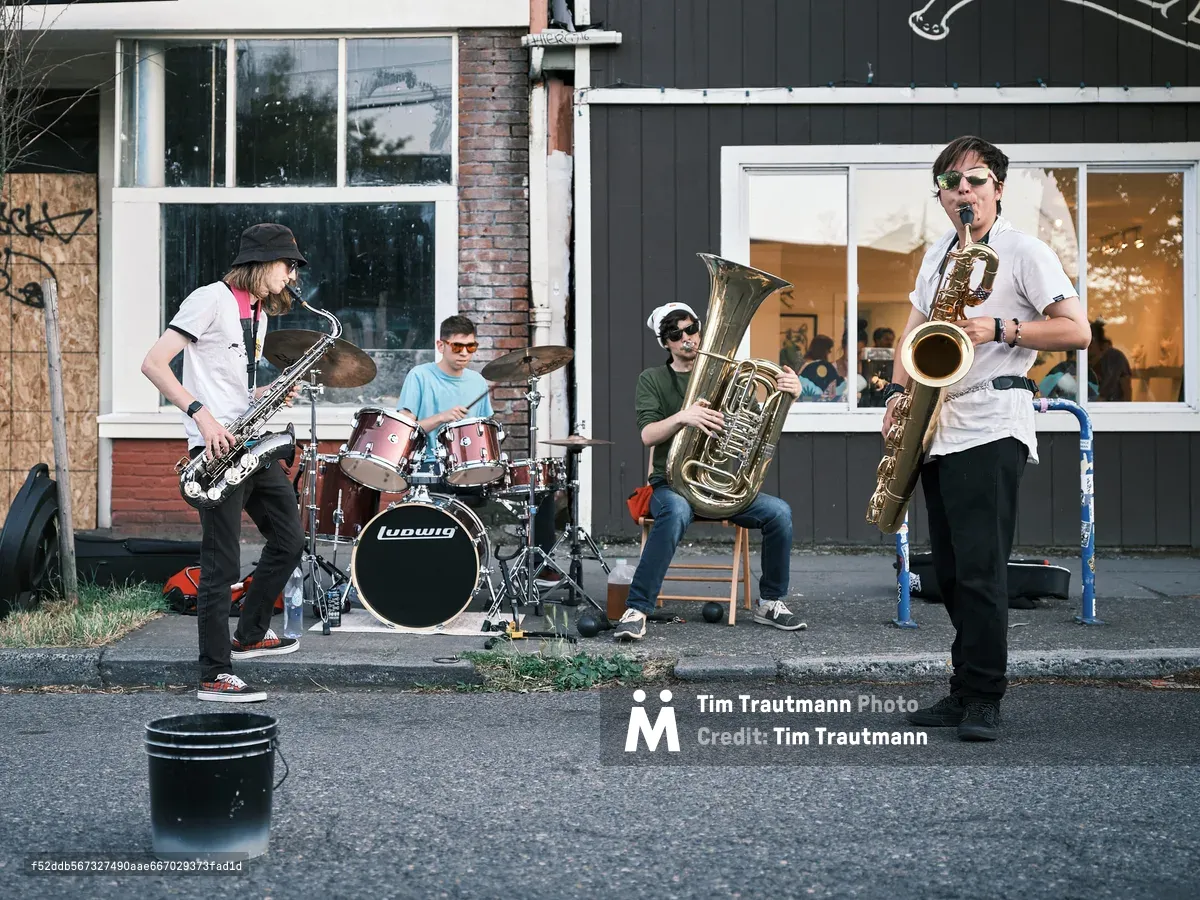 Four young musicians perform as a street band on Alberta Street during Last Thursday in Portland, Oregon. From left to right, a saxophonist in a bucket hat and white t-shirt plays tenor saxophone, a drummer in a light blue t-shirt plays a red Ludwig kit, a tuba player in a green shirt and sunglasses sits on a stool, and a baritone saxophonist in a white shirt and sunglasses plays in the foreground. A black tip bucket sits on the street in front of them. Storefronts and a gallery with people visible inside form the backdrop.
