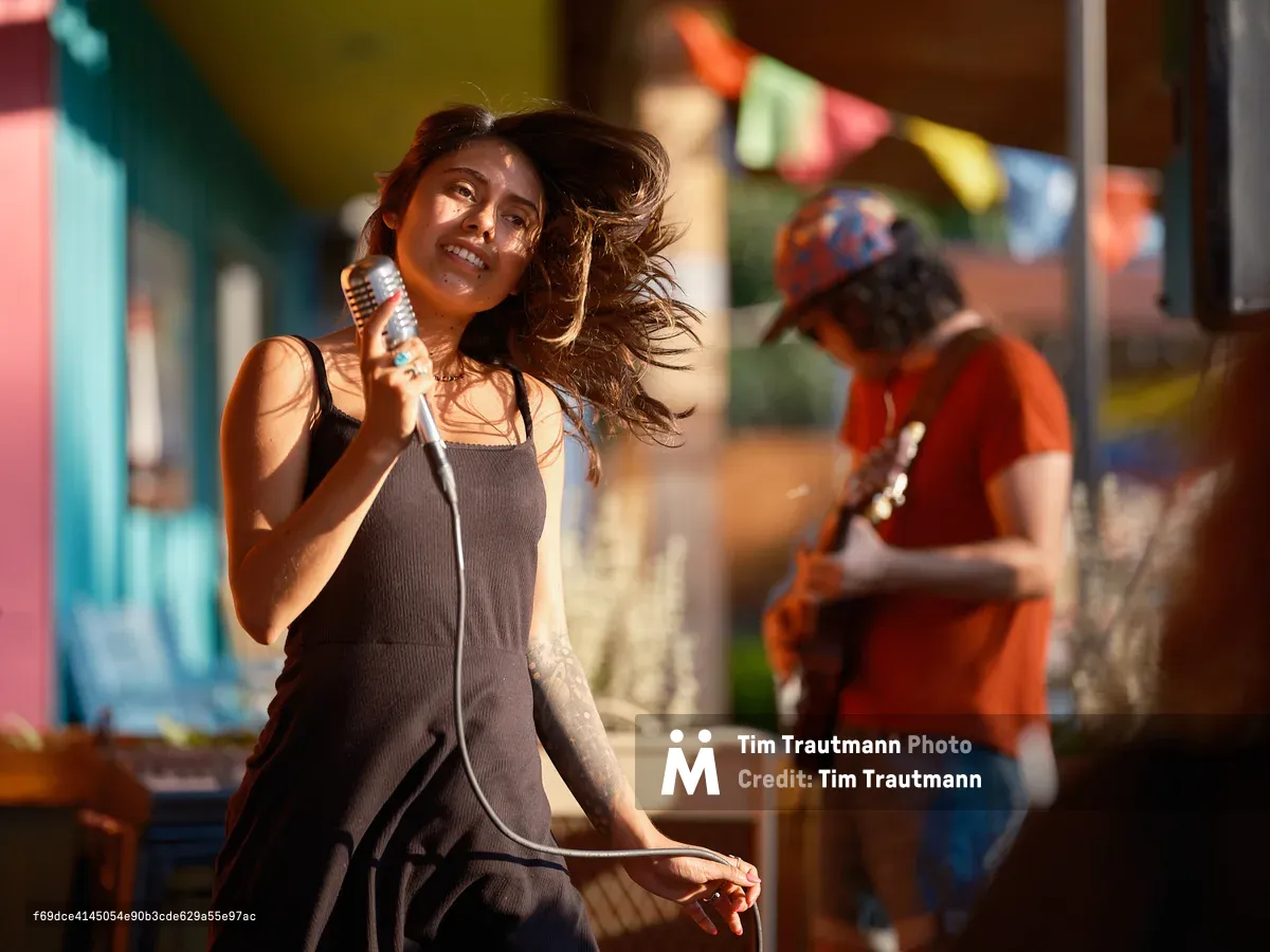 A singer performs outdoors at Portland Mercado in warm golden light, her long dark hair sweeping dramatically across her face as she moves. She holds a vintage-style silver microphone, wears a dark grey slip dress, and has a tattoo on her forearm. She smiles joyfully mid-performance. A guitarist in a red shirt and colorful cap plays in the blurred background, with the Mercado's brightly colored buildings and festive bunting visible behind them.