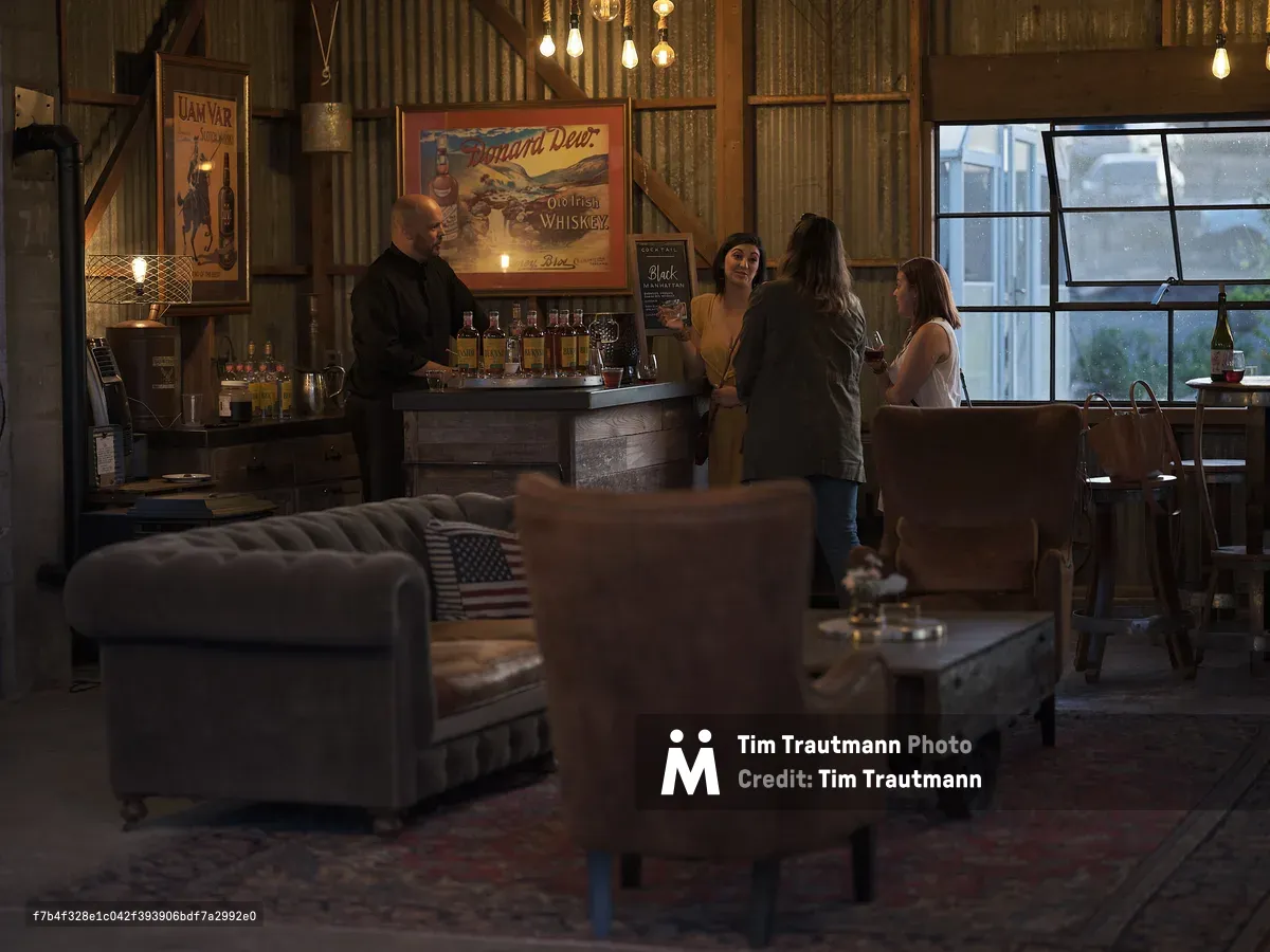 The warmly lit interior of the cocktail bar at Abbey Road Farm in Carlton, Oregon. A bartender in a black shirt stands behind a rustic wooden bar stocked with bottles of whiskey, as three women chat and enjoy drinks. Vintage whiskey advertising posters, including ones for Uam Var Scotch Whisky and Dunard Dew Old Irish Whiskey, hang on corrugated metal walls. Edison bulbs dangle from the ceiling, and the eclectic lounge area in the foreground features tufted velvet sofas, worn leather armchairs, and a Persian rug. A large industrial window lets in soft evening light on the right.